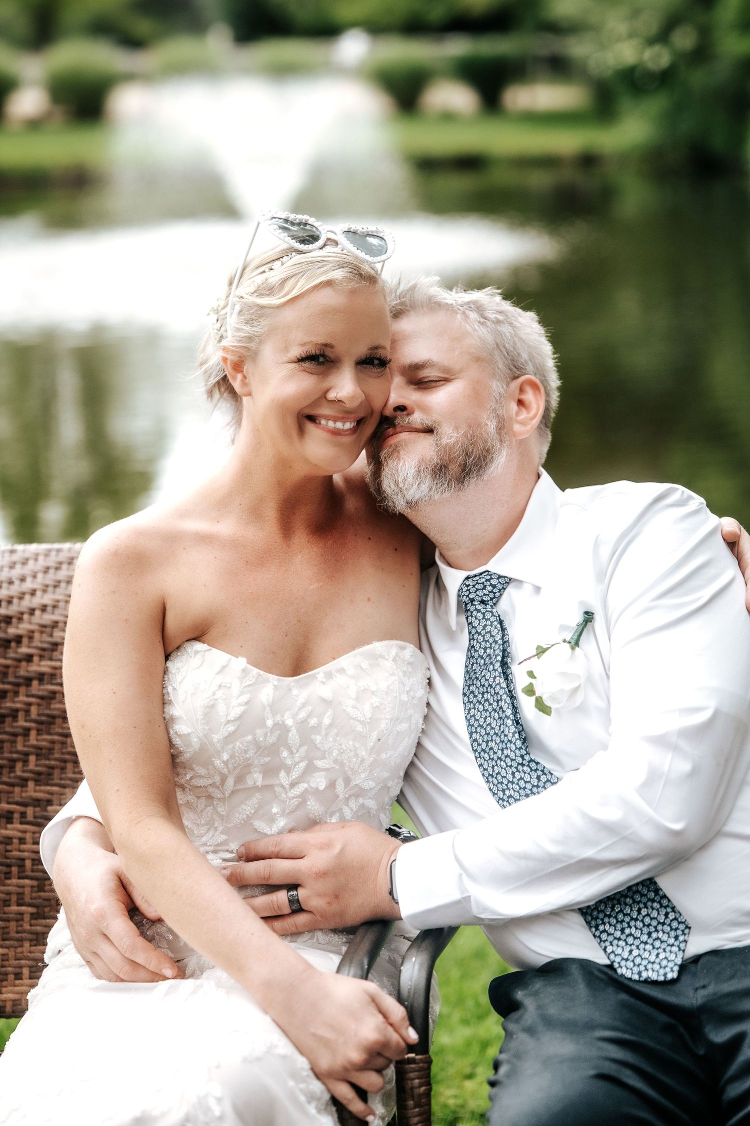 A bride and groom share a tender moment seated outdoors near a garden fountain on their wedding day.