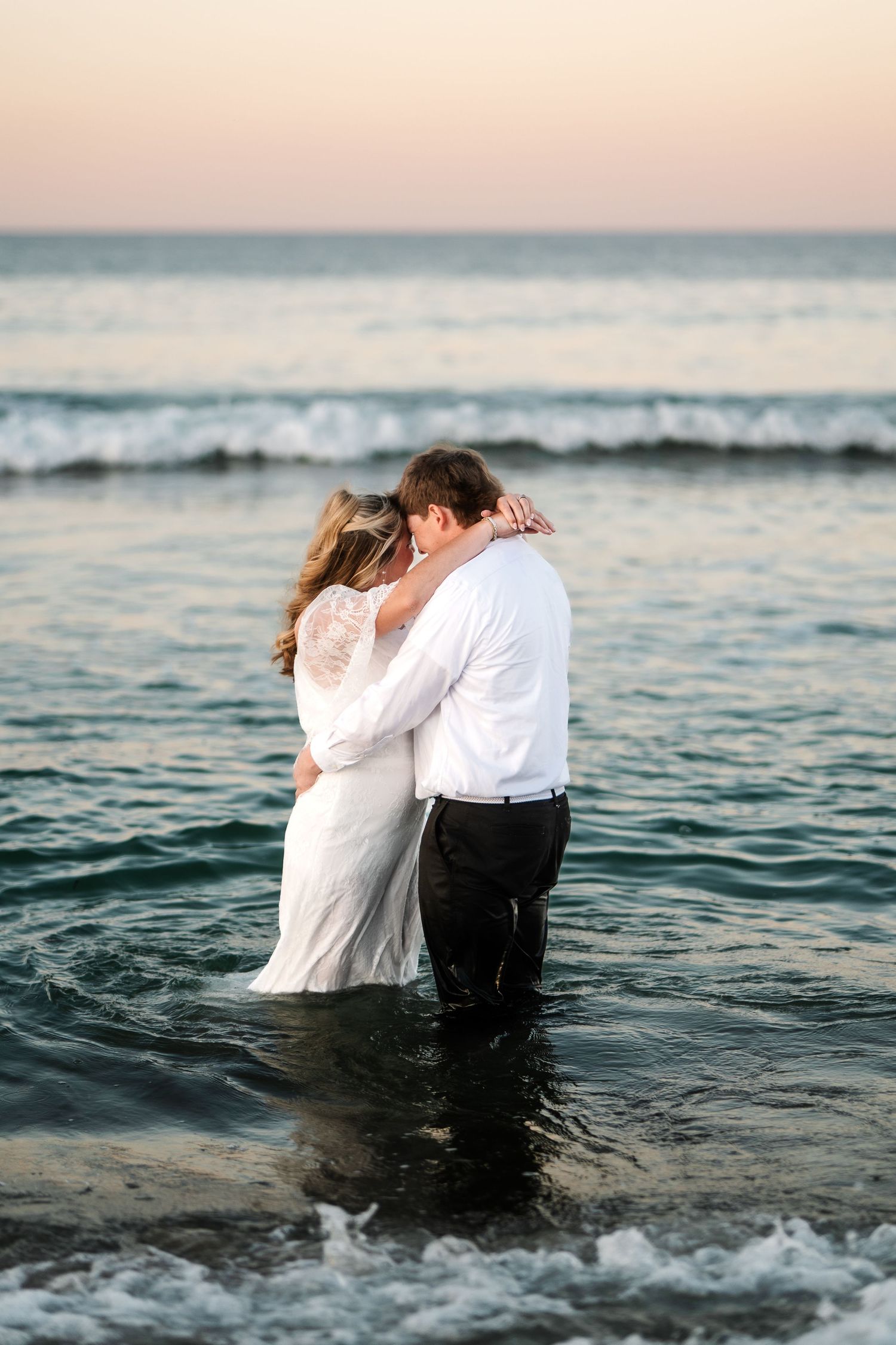 Bride and groom sharing intimate embrace standing in ocean waves at peaceful pastel sunset beach.