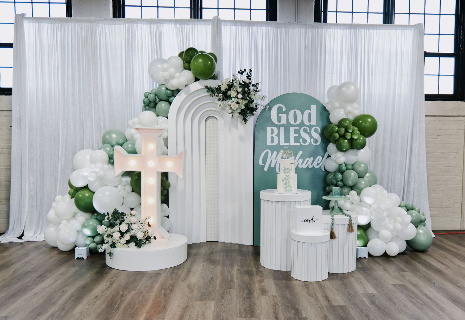 God Bless Michael christening display with white arch backdrop, illuminated cross, and balloon garland.