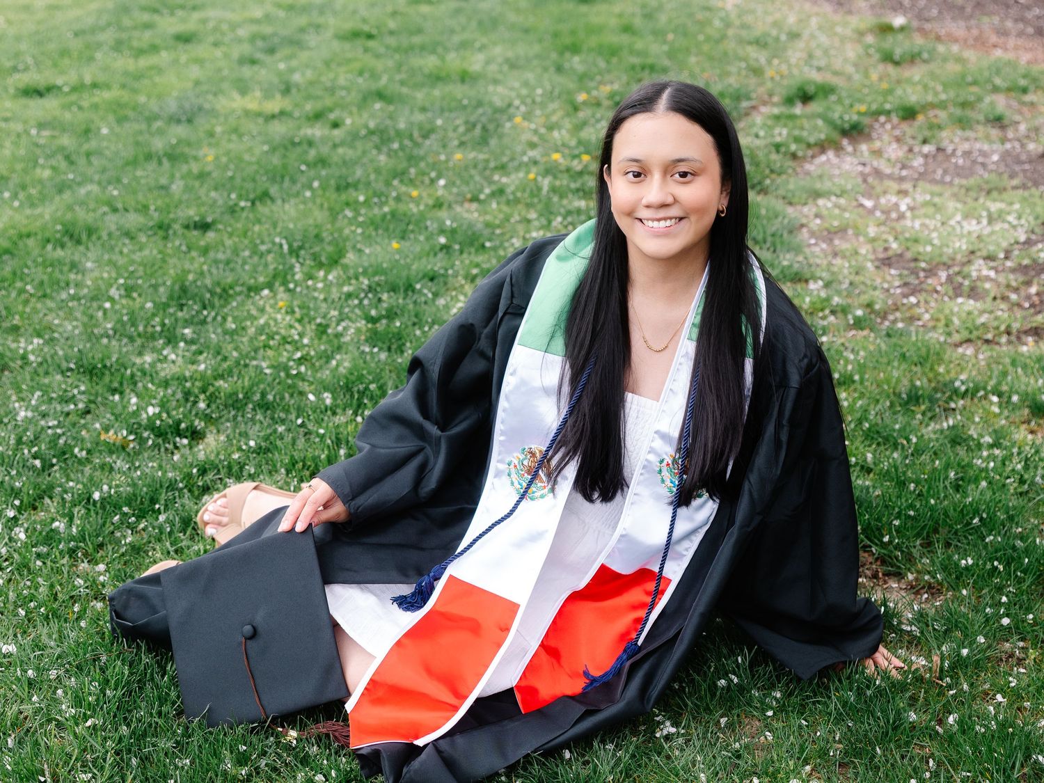 Smiling graduate in black cap and gown with Mexican flag draped over shoulders, sitting on green grass outdoors.
