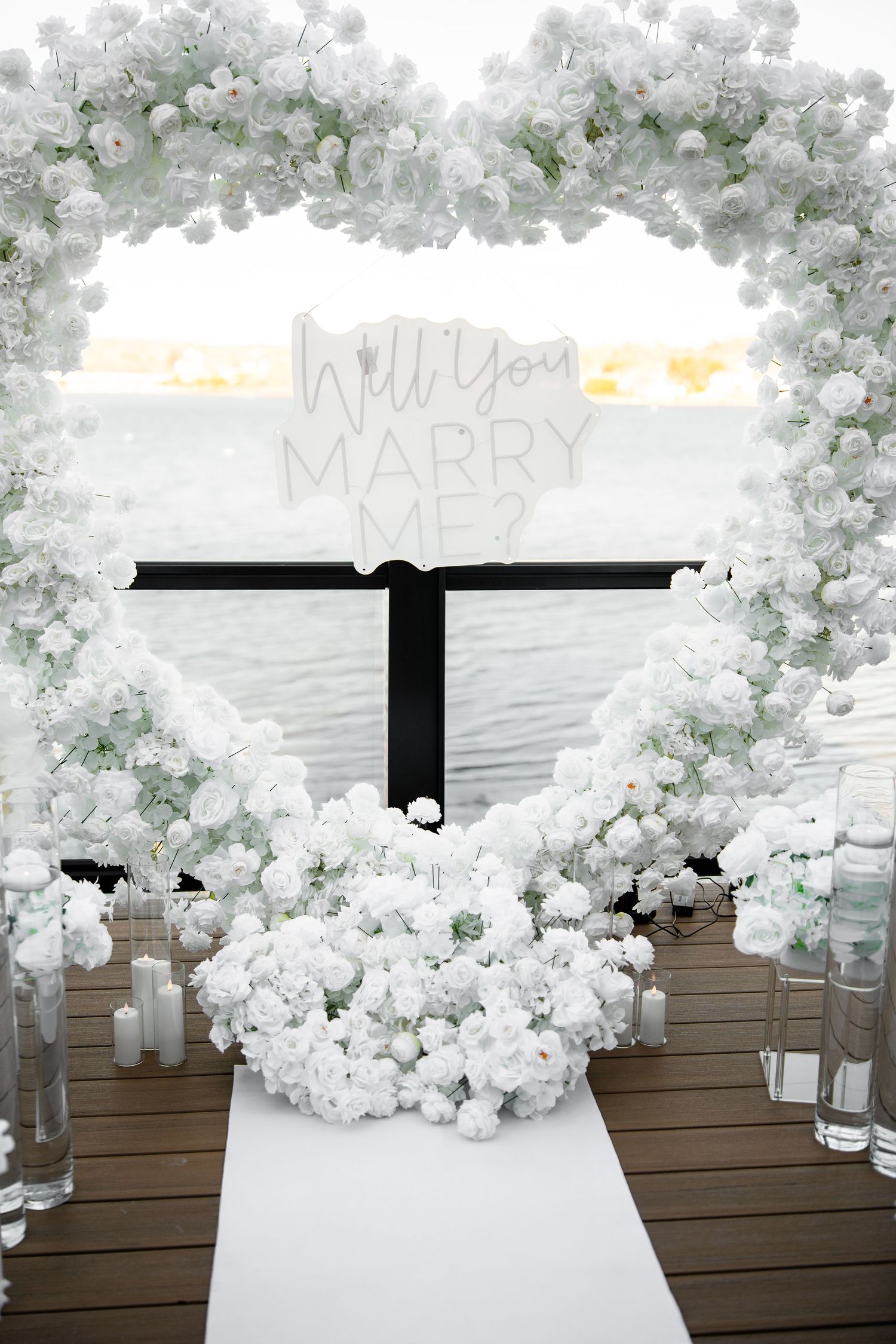 White floral arch with “Will You Marry Me?” sign over waterfront deck, surrounded by white roses and candles.