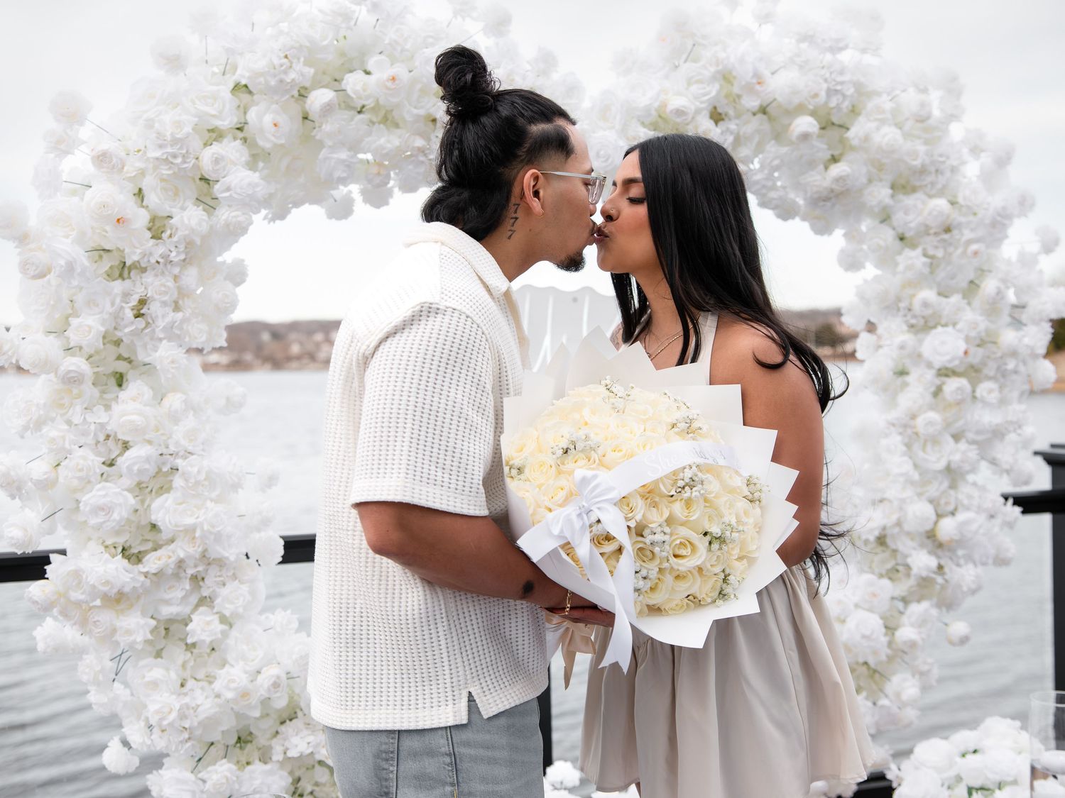 Couple kissing under a white floral arch while woman holds a bouquet of white roses outdoors.