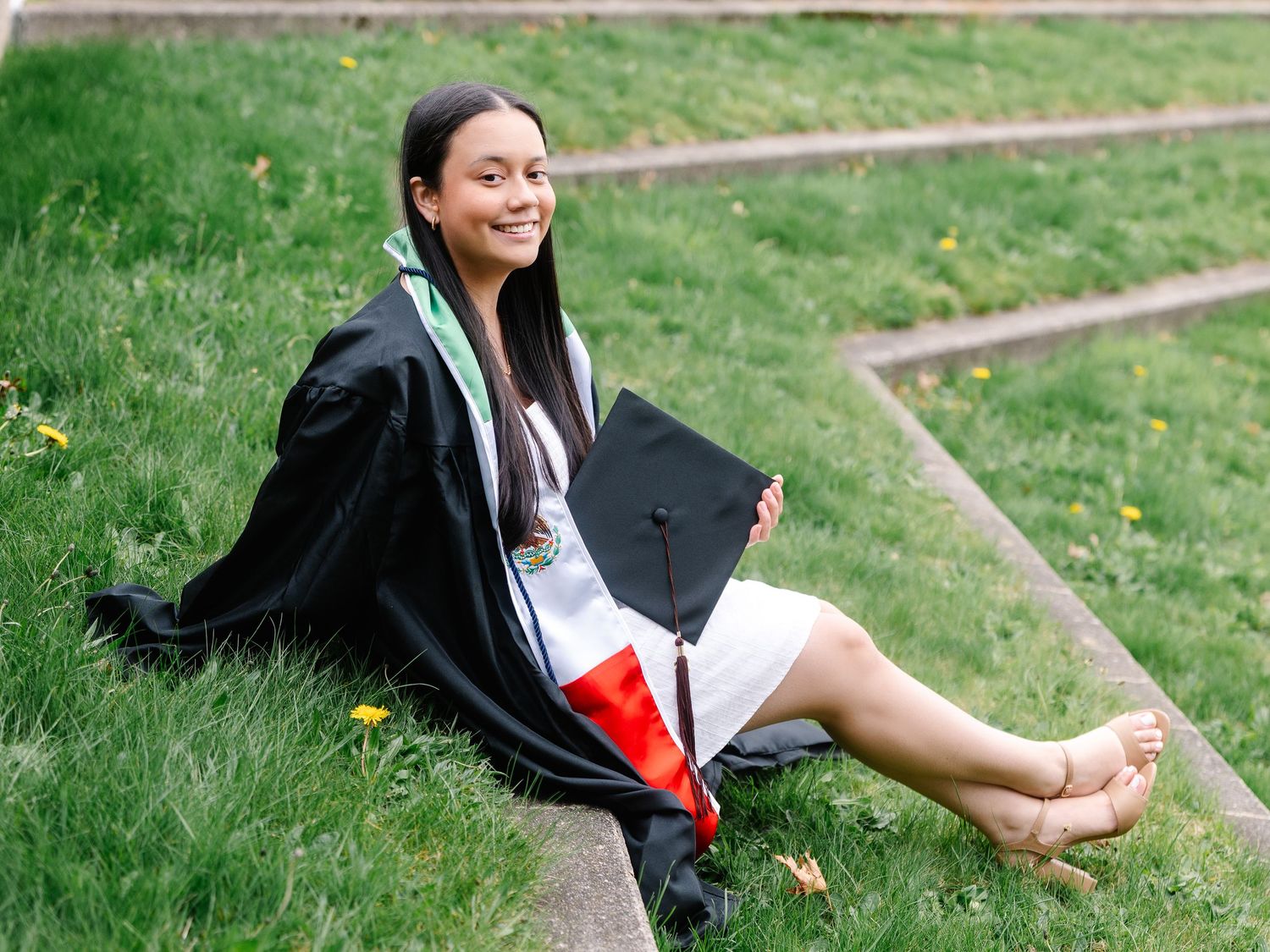 Young woman in black graduation gown and cap sitting on grass, smiling, wearing a medal and colorful sash.