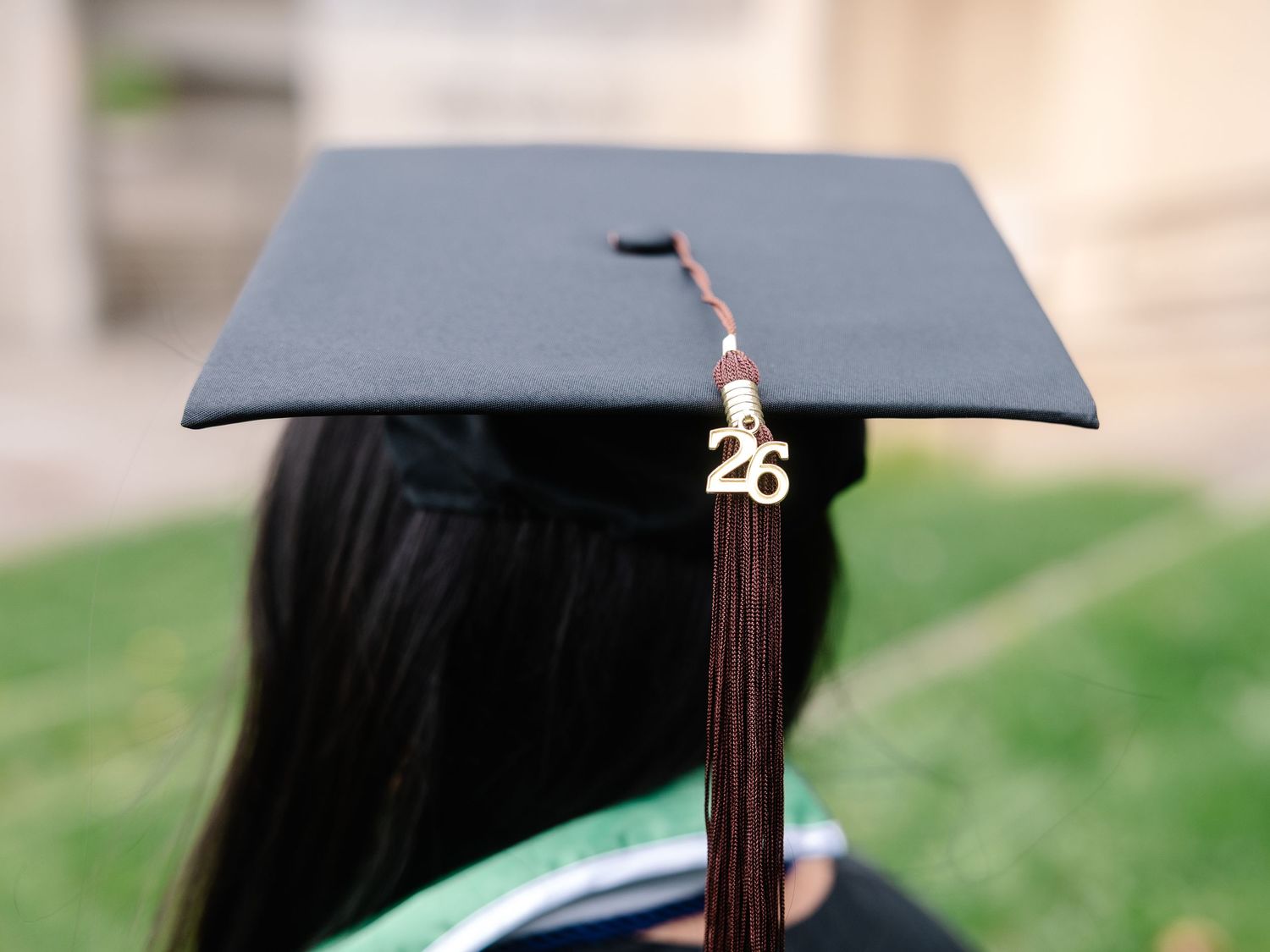 Graduate wearing black mortarboard cap with gold 26 tassel charm, viewed from behind at graduation ceremony.