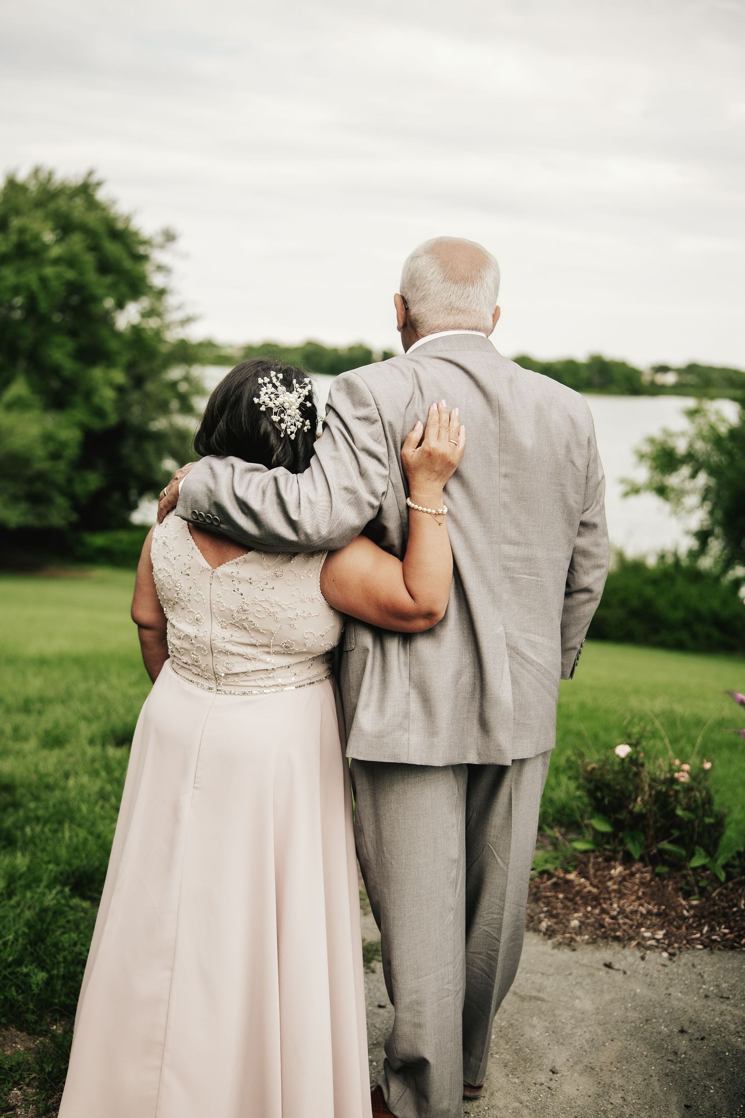 Bride and groom walking arm in arm outdoors, viewed from behind, wearing wedding attire near a scenic waterfront.