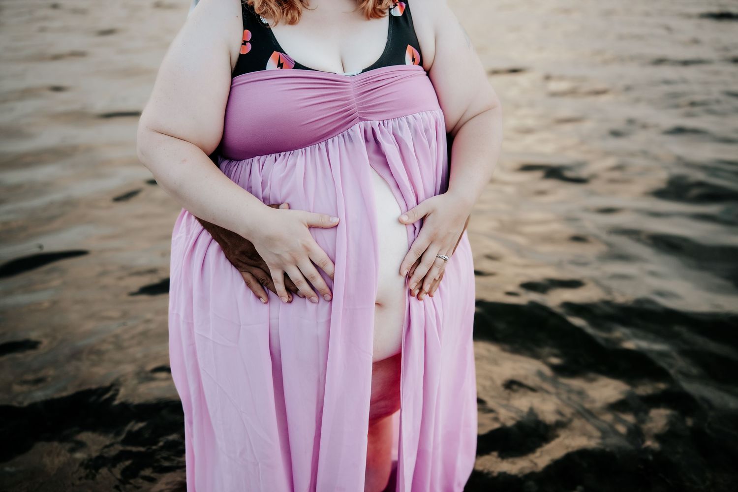 Pregnant woman in pink flowing maternity gown standing by water, cradling her baby bump at golden hour.
