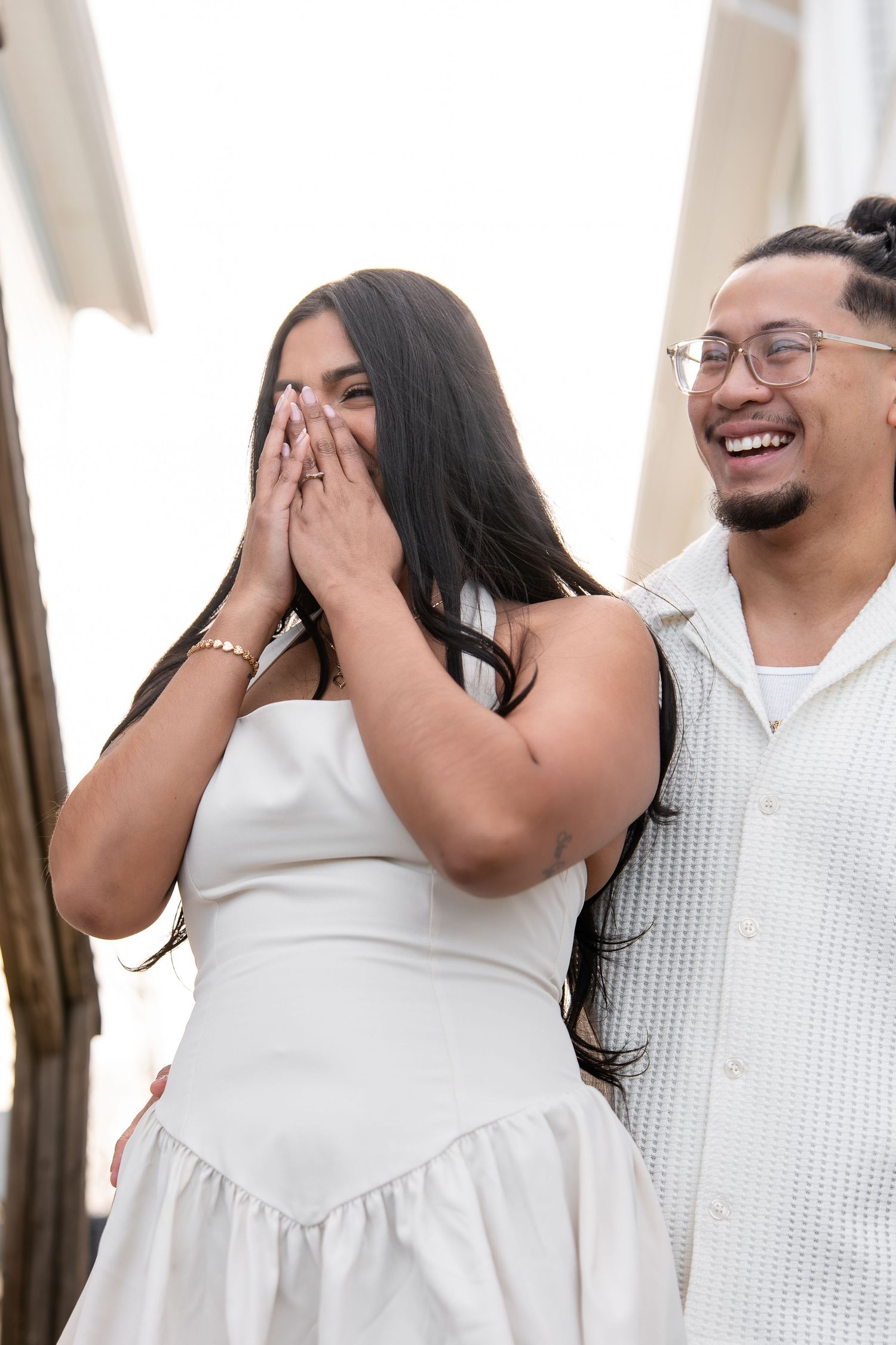 Couple laughing joyfully outdoors, woman in white dress covering her mouth, man in white shirt smiling beside her.