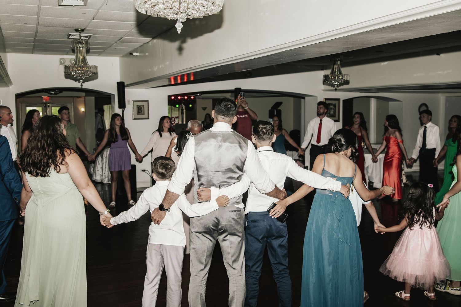 Guests holding hands in a circle dance at a wedding reception in an elegant banquet hall.