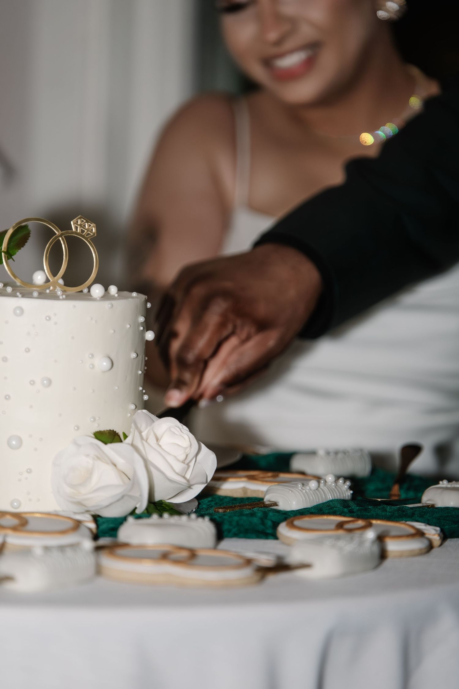 Bride and groom cutting a white wedding cake topped with gold rings, surrounded by decorated cookies on a green table.
