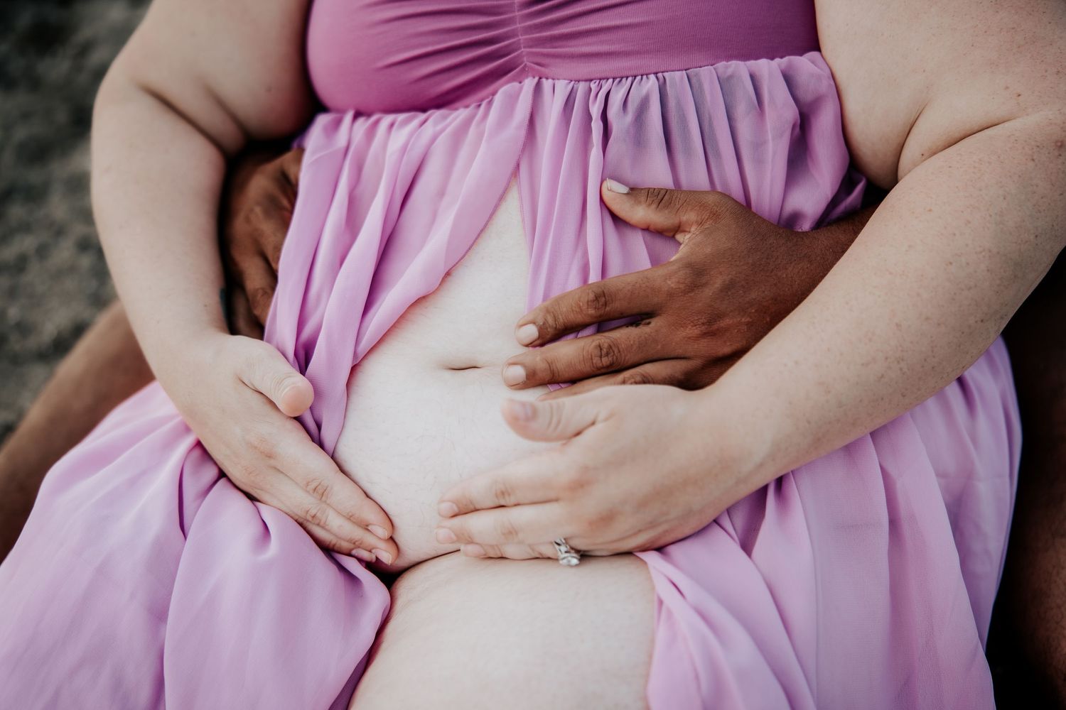 Pregnant woman in lavender dress with two partners hands cradling her bare baby bump outdoors.