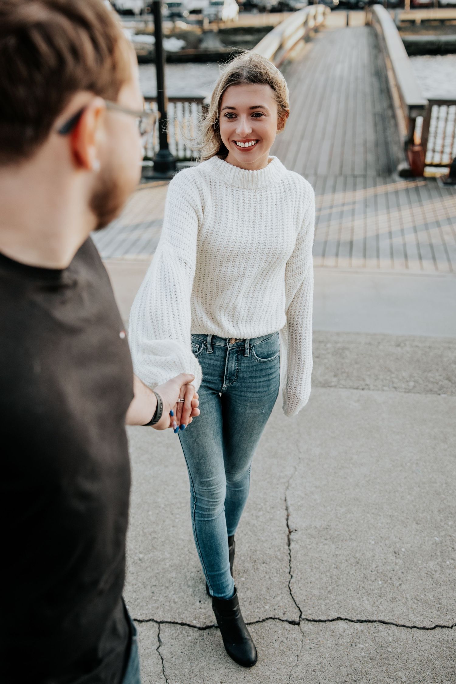 Smiling woman in white knit sweater and jeans holds mans hand near a waterfront boardwalk.