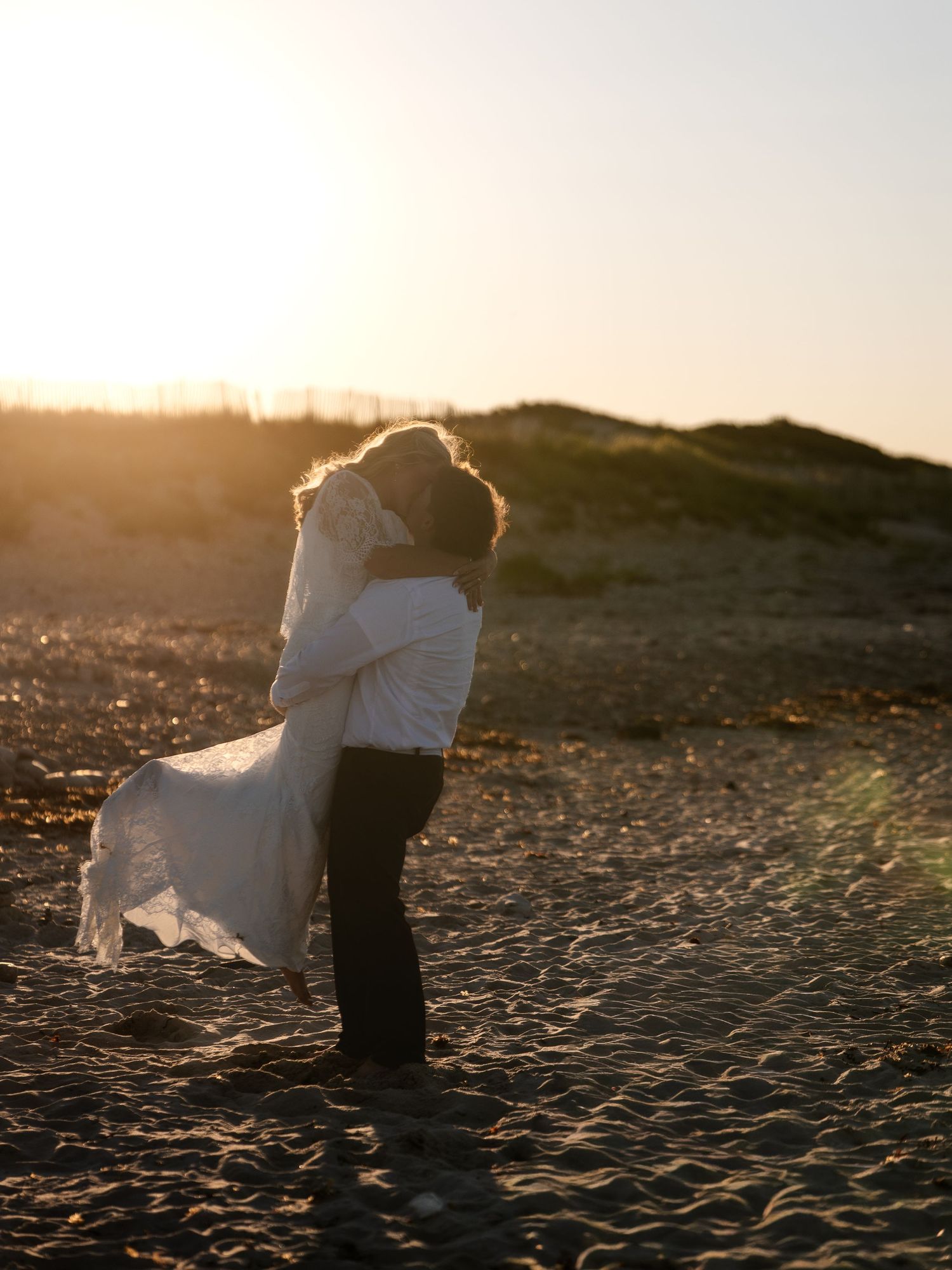 Groom lifting bride in white lace wedding dress on sandy beach during stunning golden hour sunset.