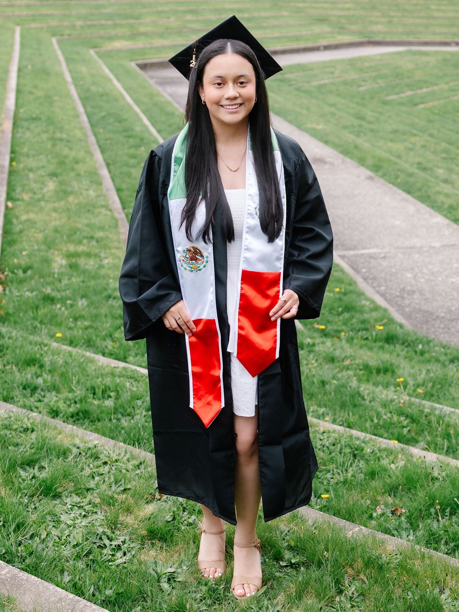 Graduate poses on grass steps wearing black gown and Mexican flag graduation stole.