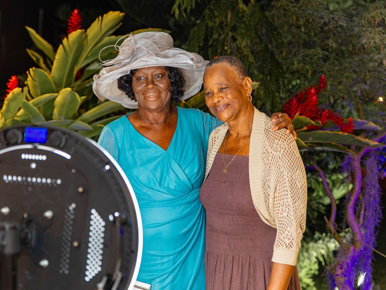 Two elegantly dressed women posing together at a formal outdoor event surrounded by tropical flowers.