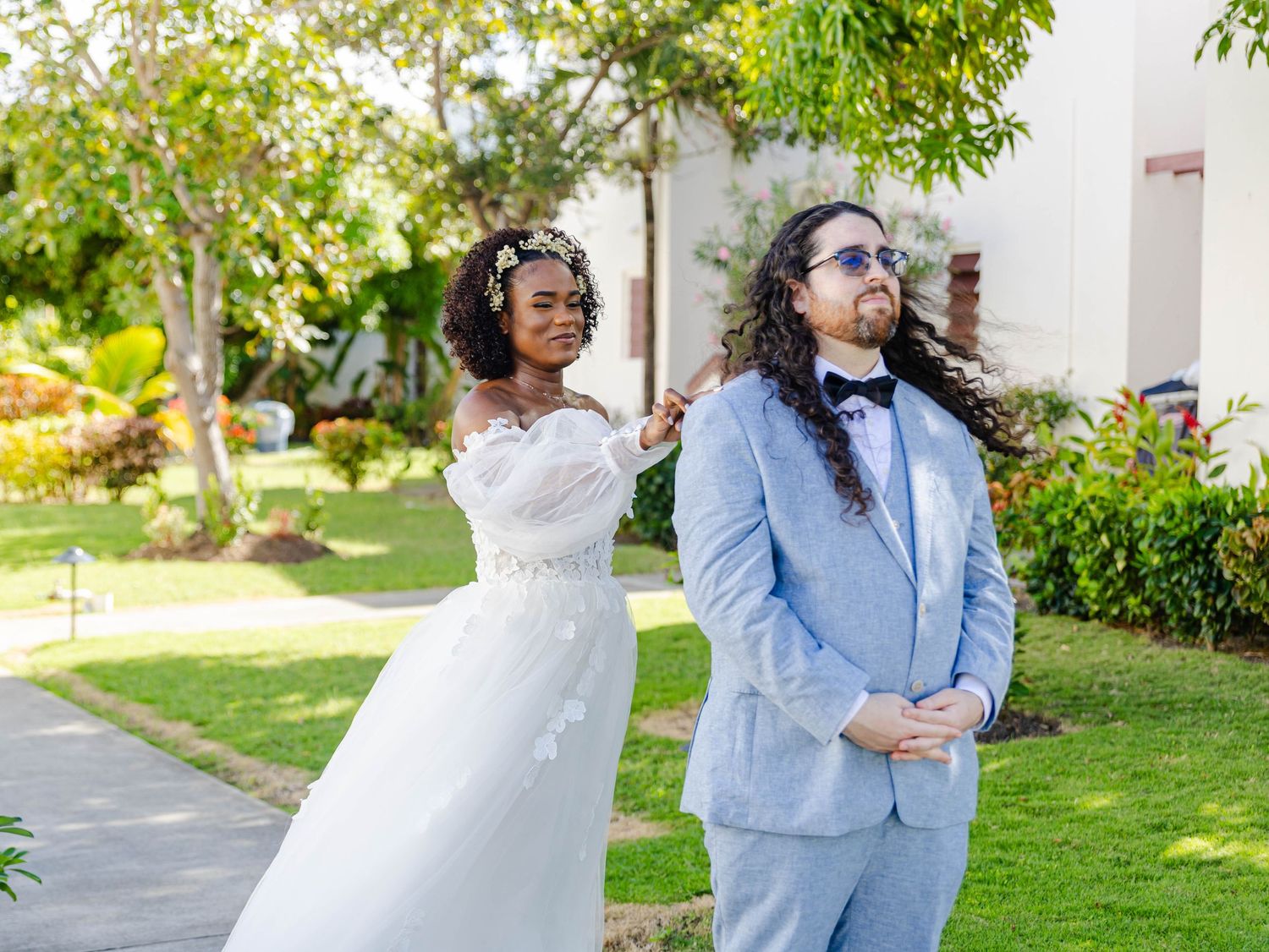 Bride in white gown taps groom's shoulder during first look, groom in light blue suit with sunglasses.