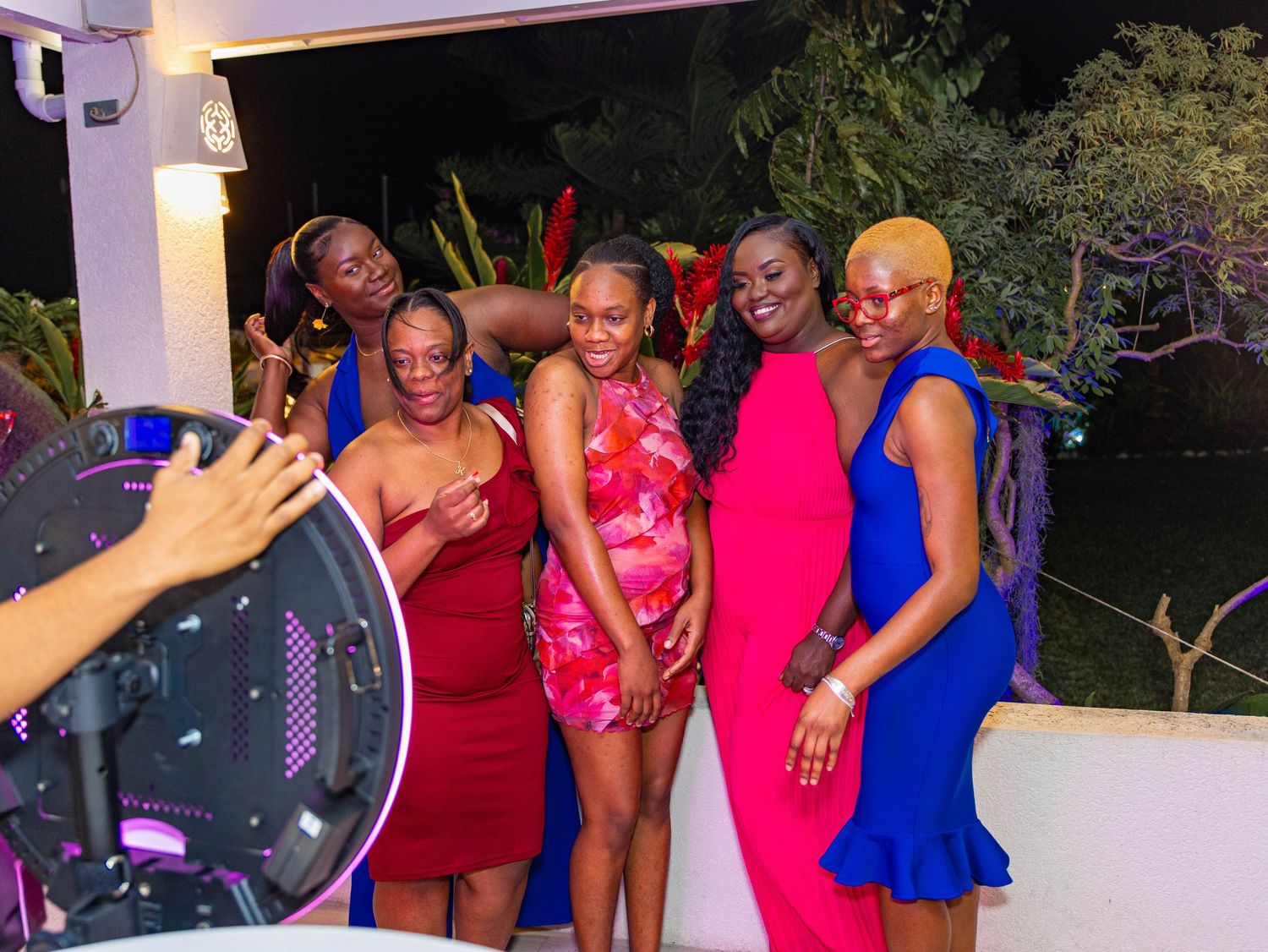 Five women posing together at an outdoor evening party, wearing colorful dresses, smiling near a DJ setup.