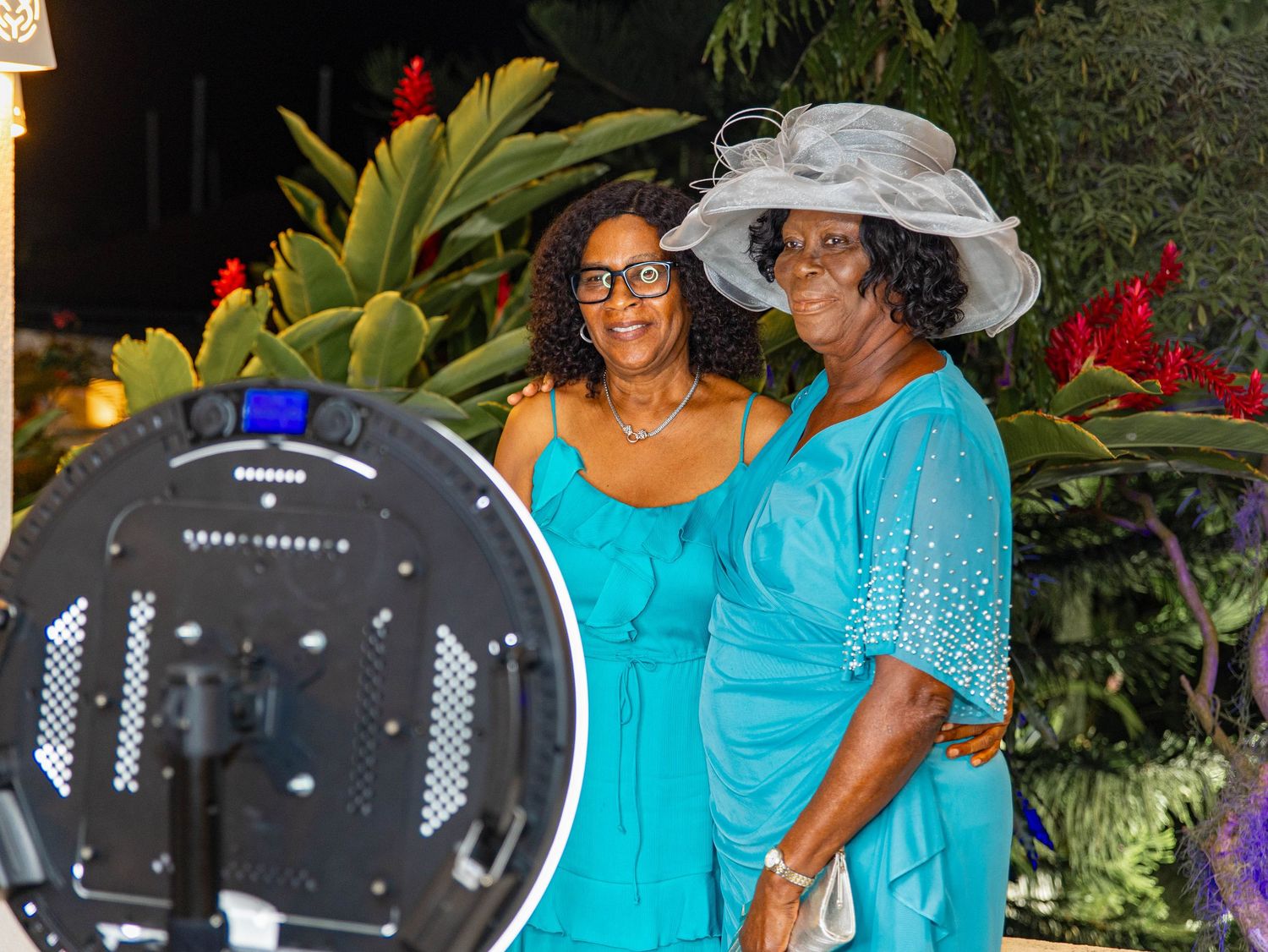Two women in teal outfits pose together outdoors at a formal event, one wearing an elegant white hat.