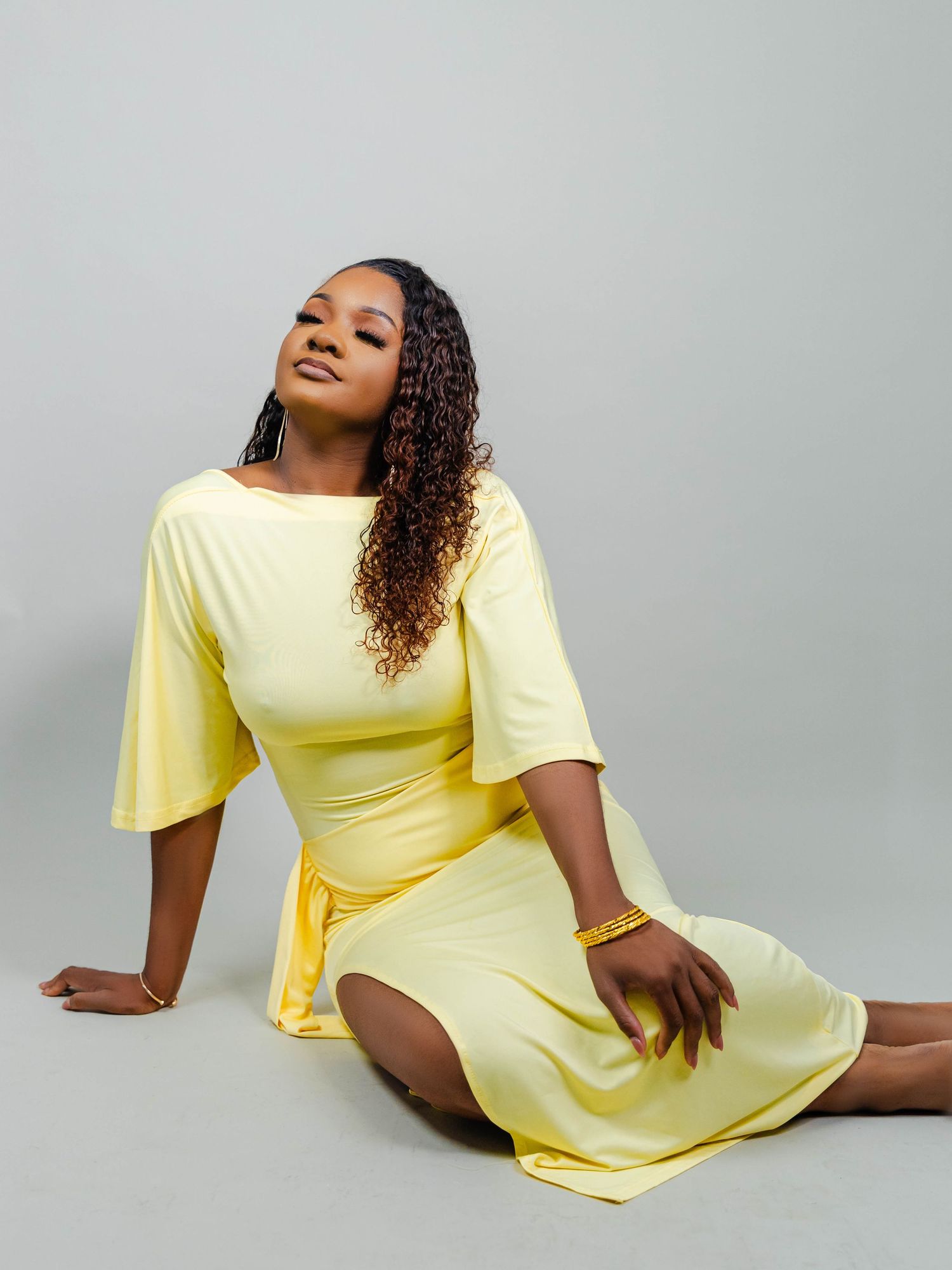 Woman in a yellow midi dress sitting gracefully on the floor, wearing gold jewelry, with curly hair against a gray background.