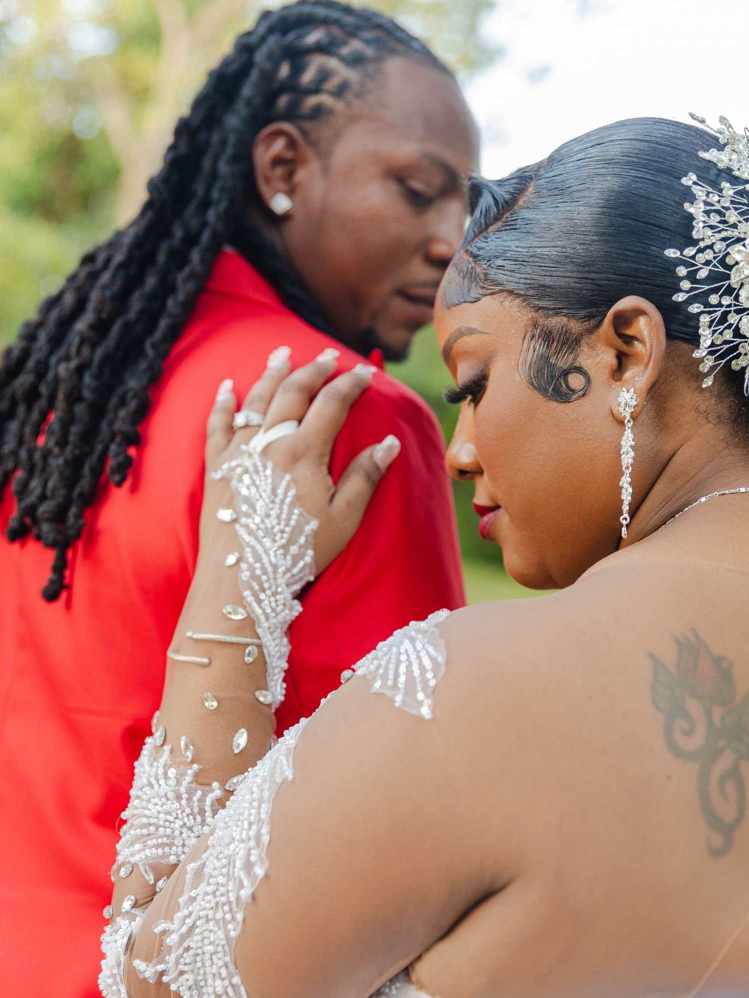 Bride in a beaded white gown with a crystal hairpiece stands close to the groom in a red suit during outdoor wedding ceremony.