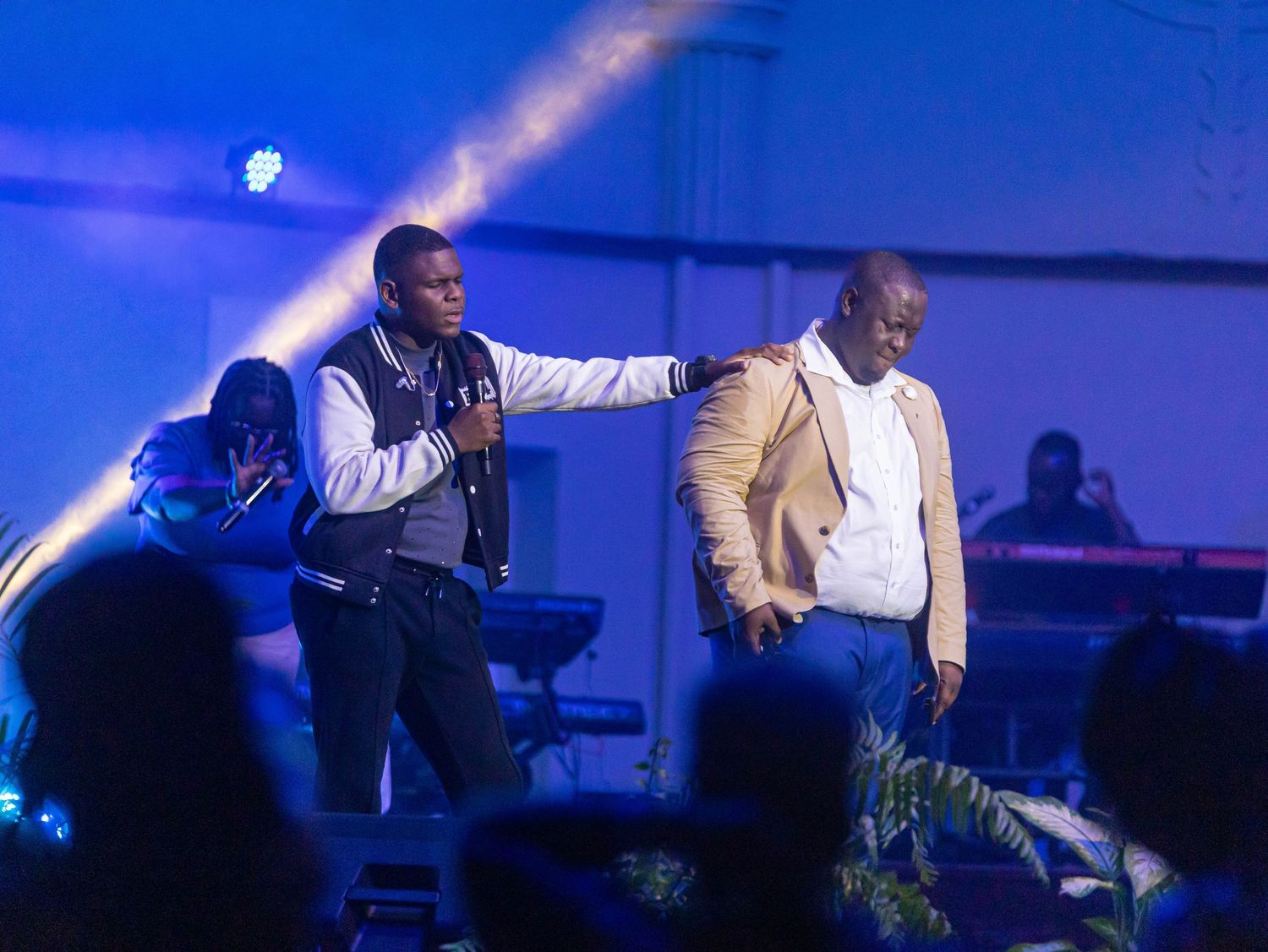 Two male performers on stage under blue lights, one holding microphone while gesturing to the other.