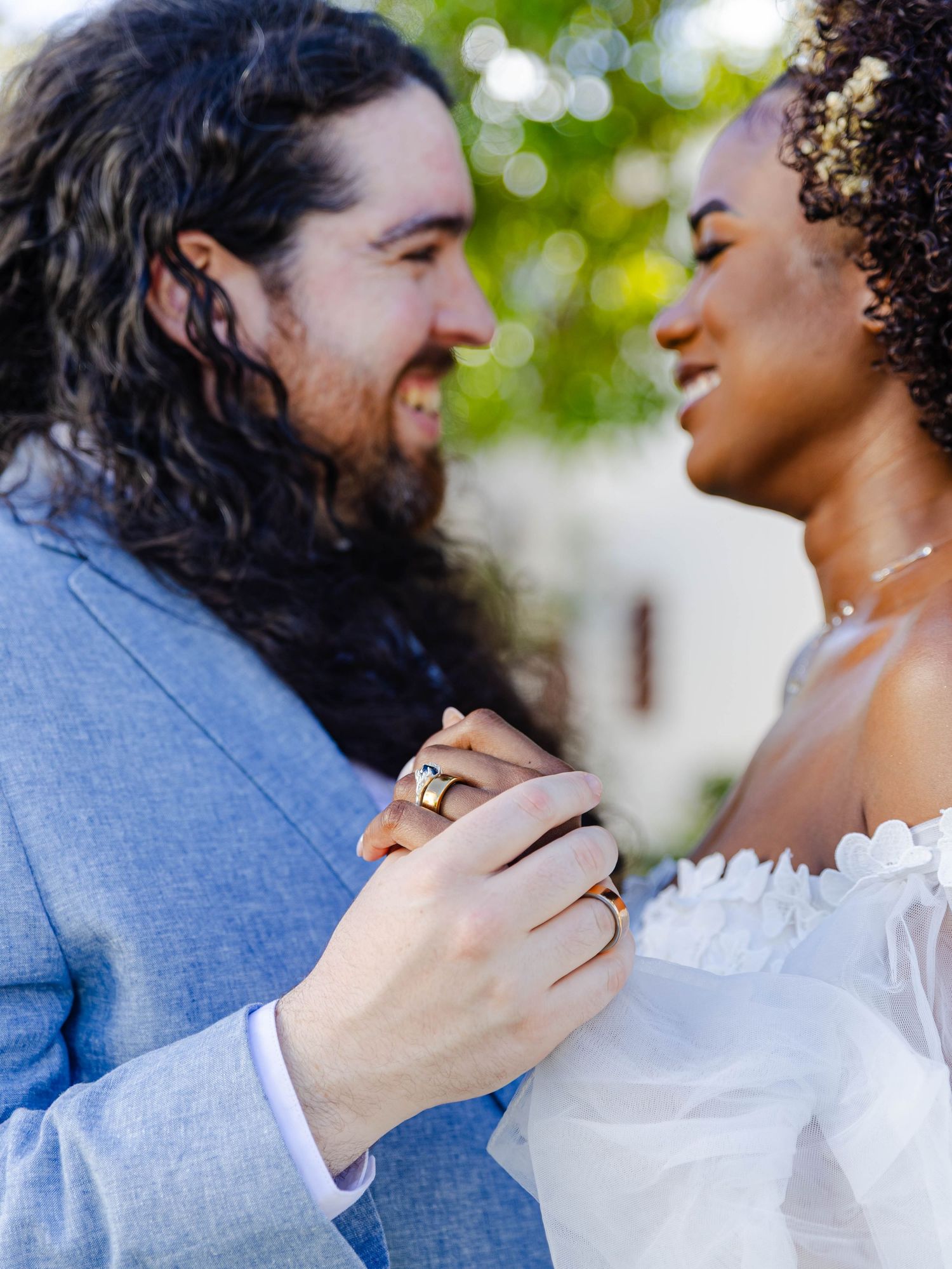 Interracial couple in wedding attire holding hands and smiling at each other outdoors.