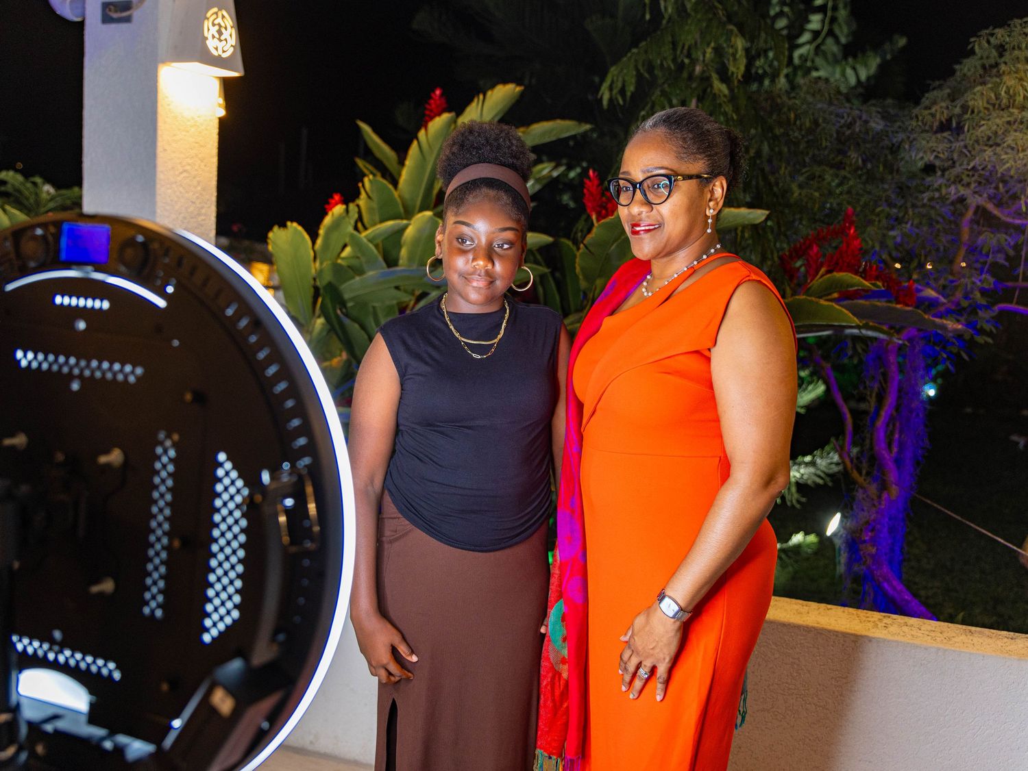 Two women posing together at an outdoor evening event, one in brown and one in orange dress, with lush greenery backdrop.
