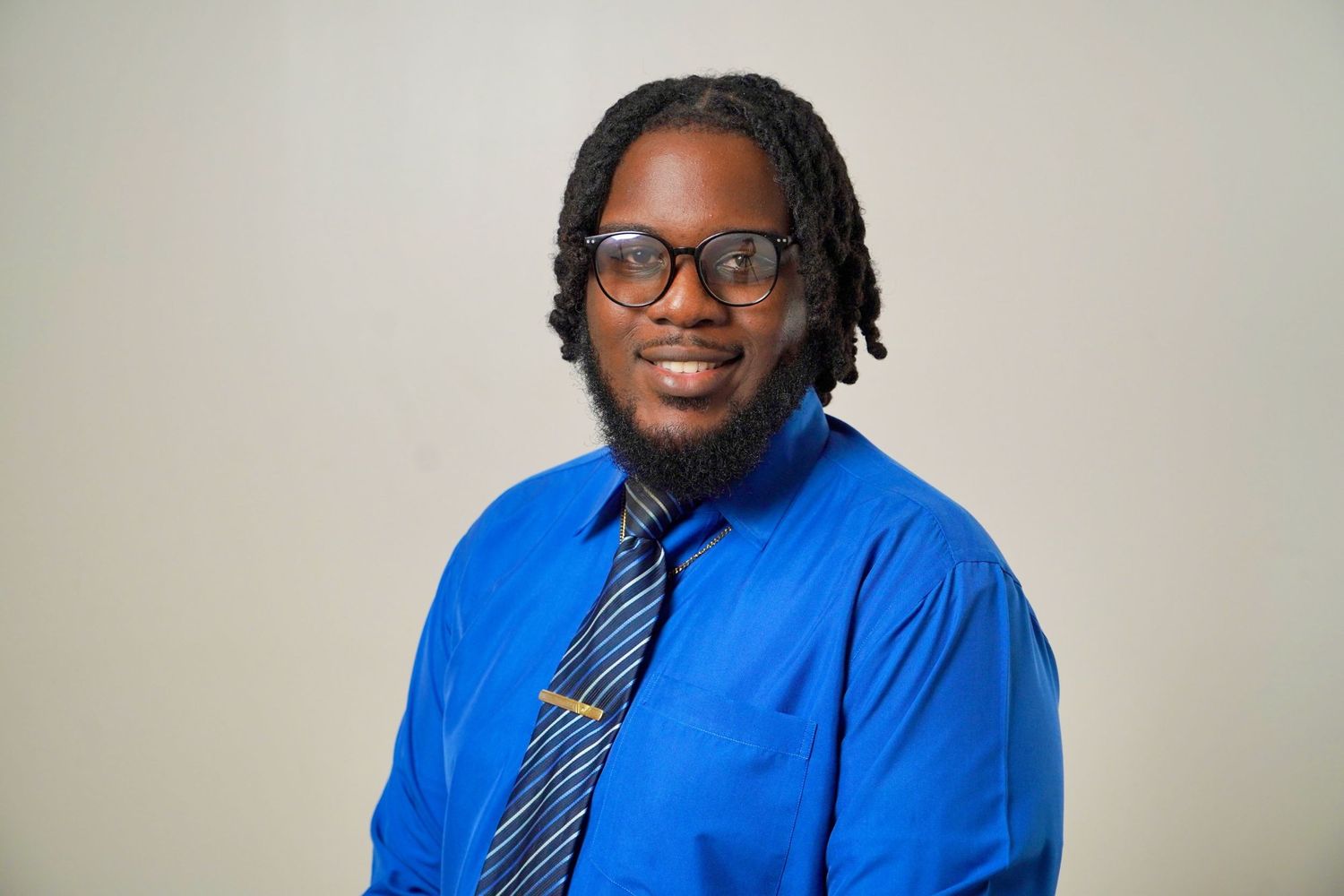 Jaleque is wearing a blue shirt and a striped tie, smiling confidently in a studio portrait.
