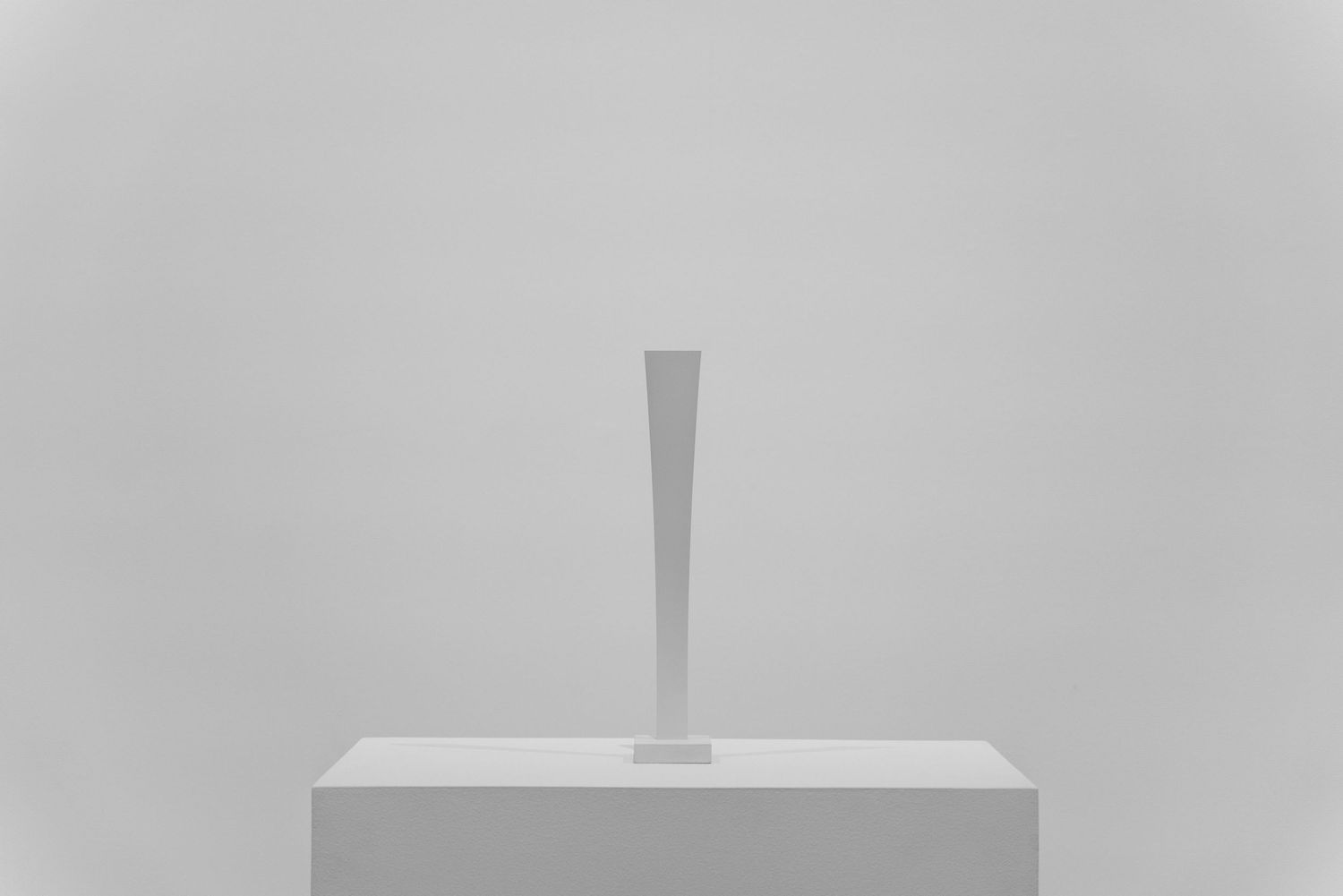 Minimalist white tapered column sculpture displayed on a white pedestal against a neutral gray background.