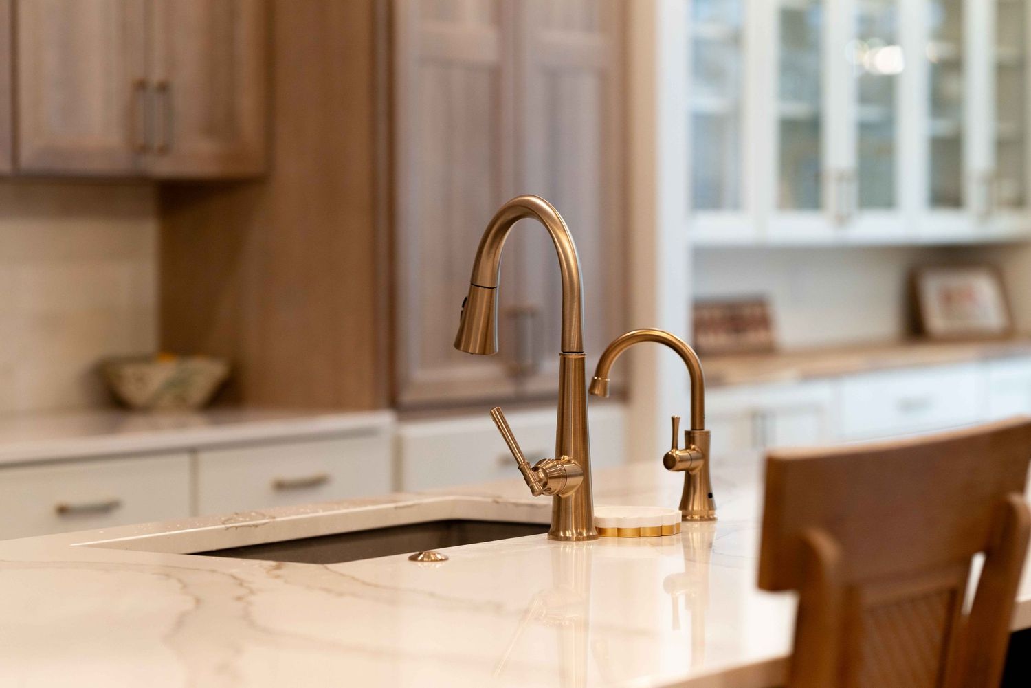 Brushed gold kitchen faucet and soap dispenser on white marble island with wood cabinetry backdrop.