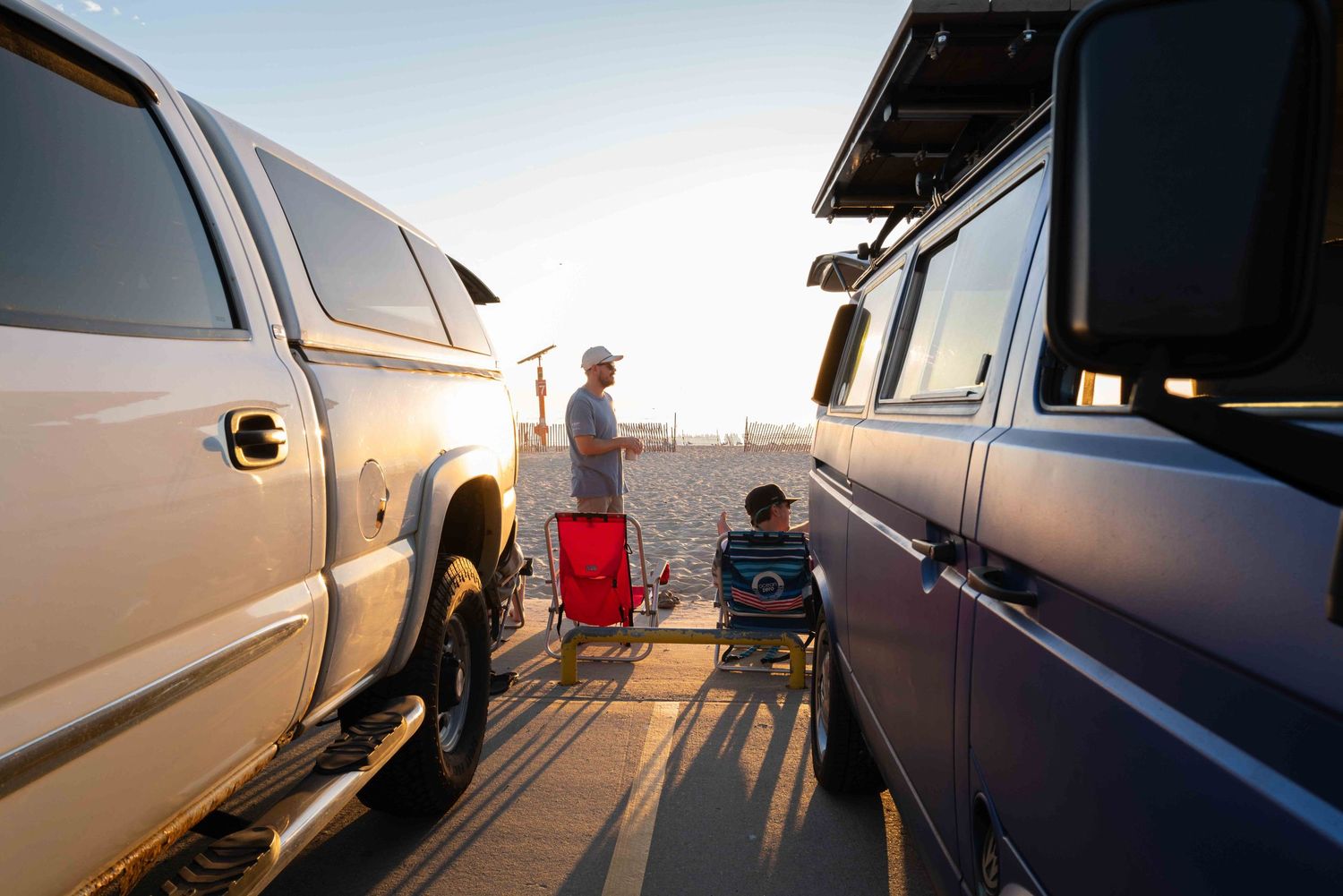 Two people relaxing in chairs between trucks on a sunny beach at sunset.