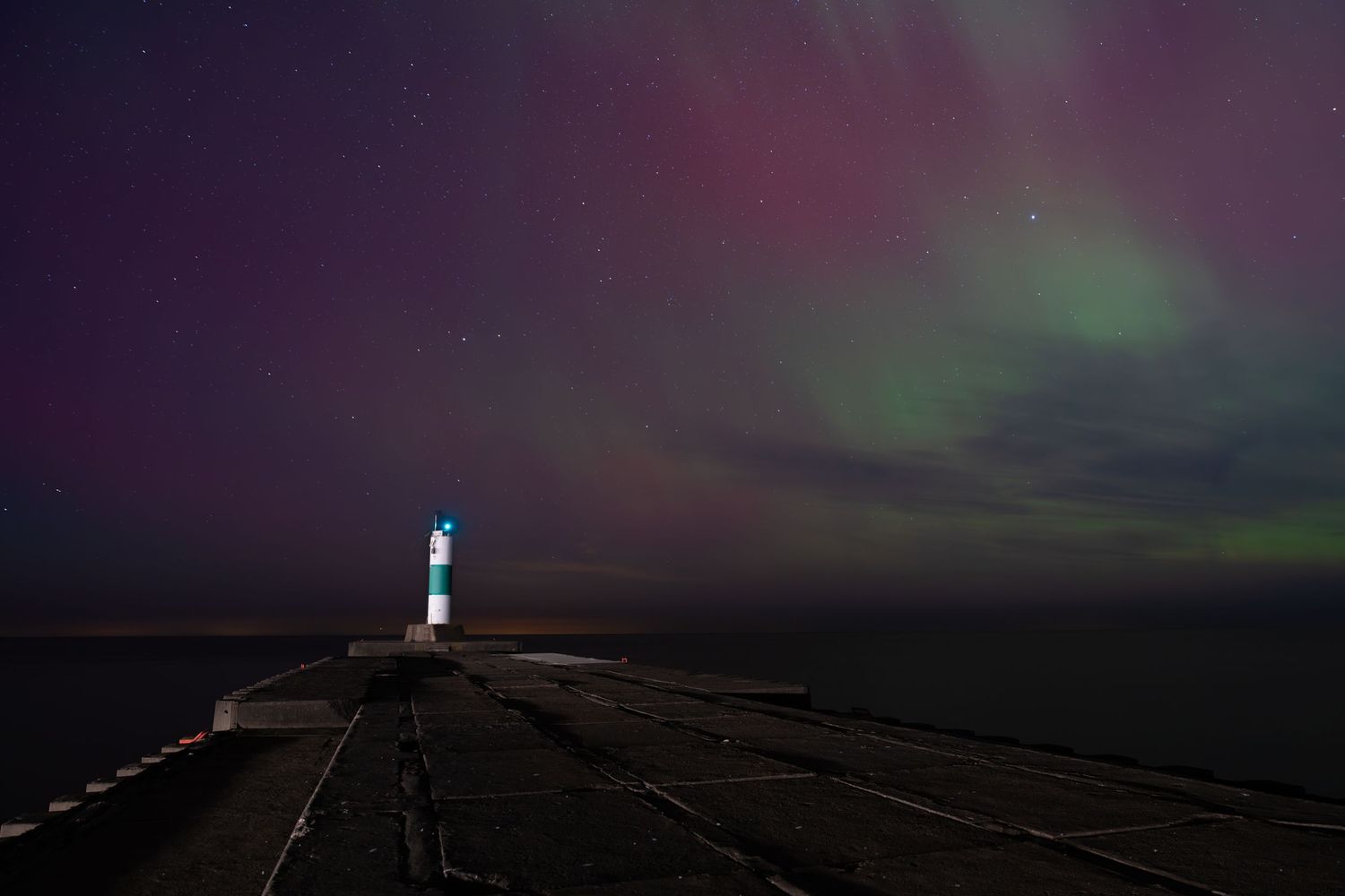 Lighthouse at end of stone pier glowing under dramatic aurora borealis with green and purple hues.