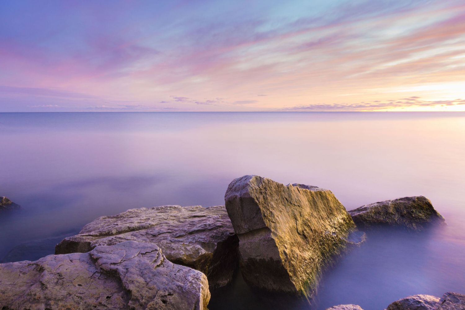 Smooth purple and pink sunset over calm ocean waters with rocky foreshore in the foreground.