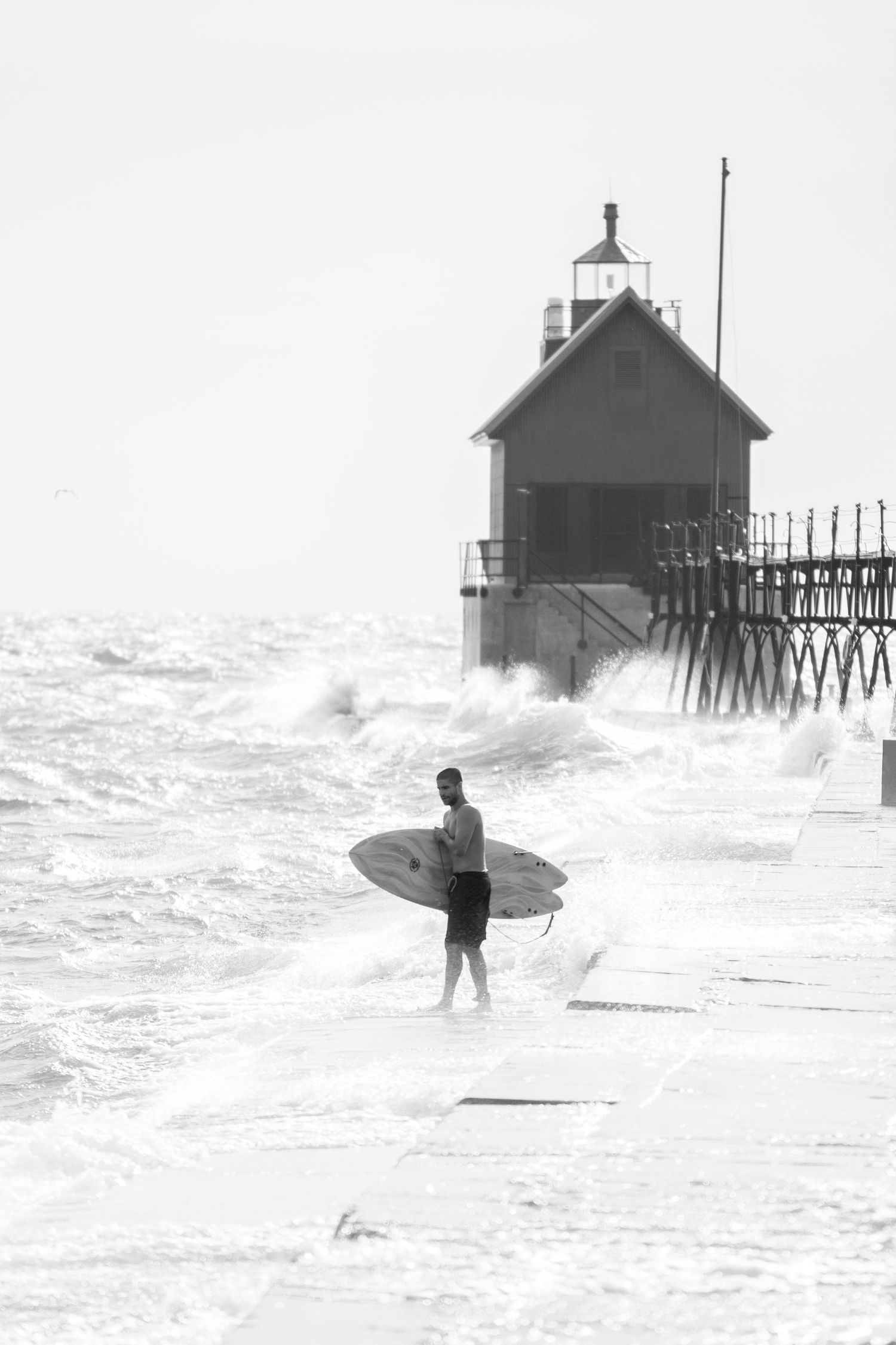 Surfer carrying board near a lighthouse pier with crashing waves in black and white.