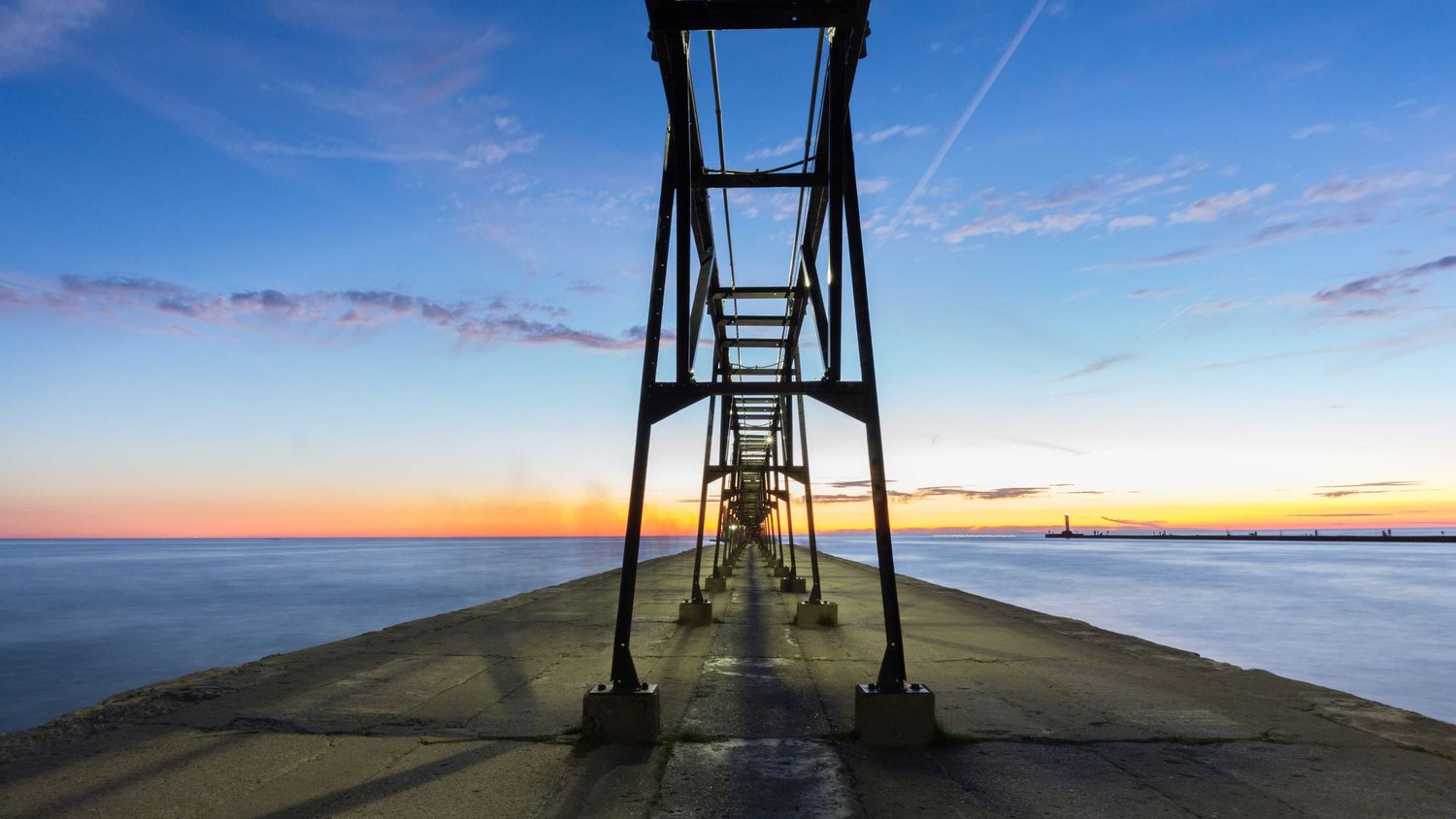 Symmetrical low-angle view of a concrete pier with metal framework extending toward a glowing sunset horizon.