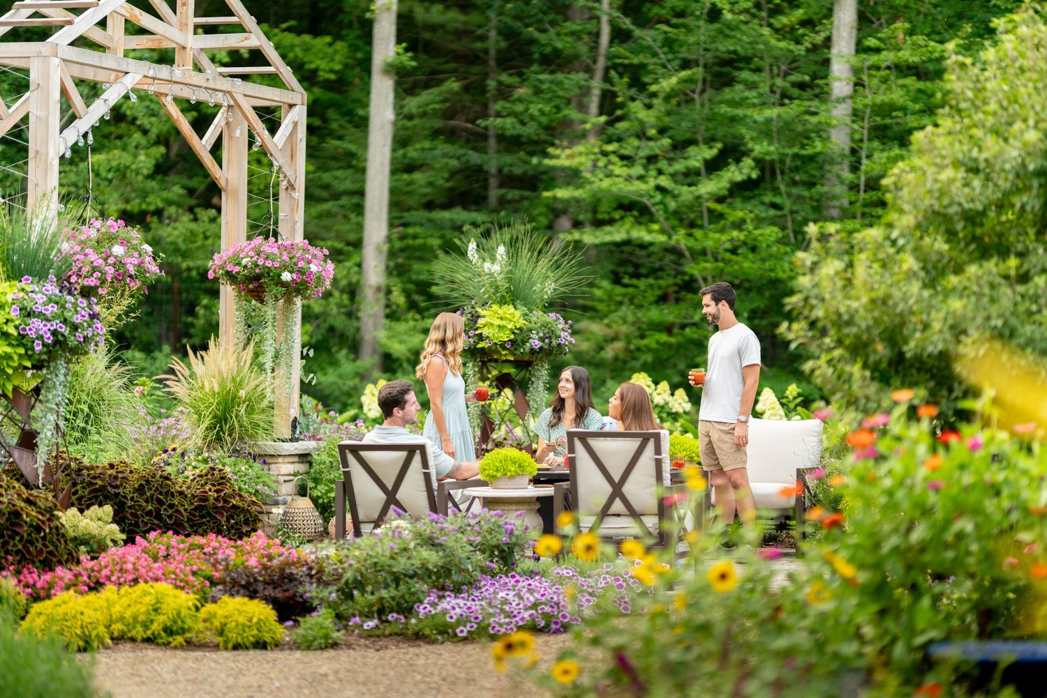 Group of people gathering around an outdoor patio table surrounded by a colorful garden landscape.