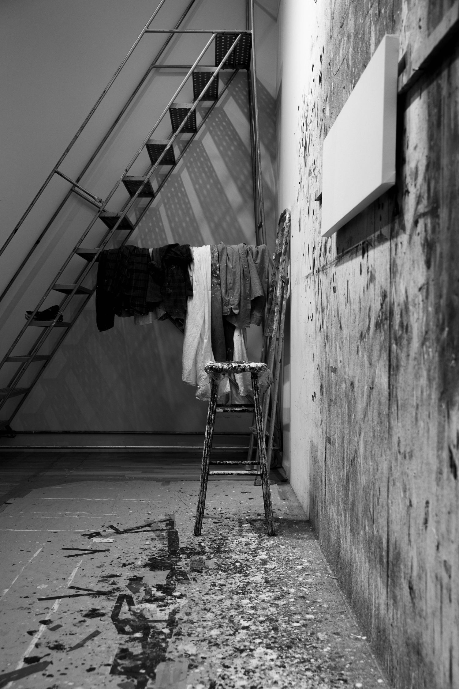 Black and white photo of abandoned room with ladder, hanging clothes, and debris on floor.