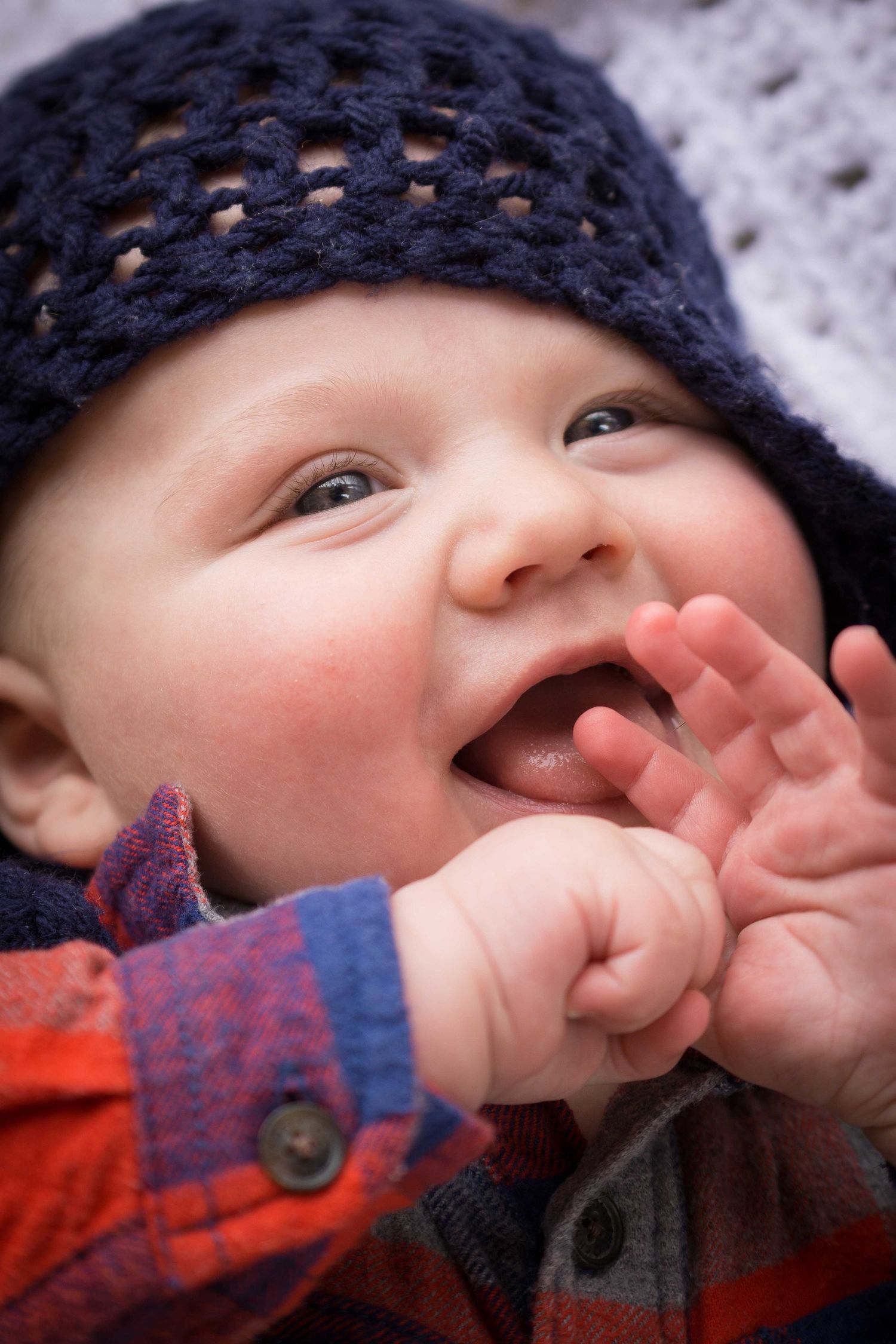 A happy baby in a navy knit hat and plaid shirt laughs with hand raised to mouth.