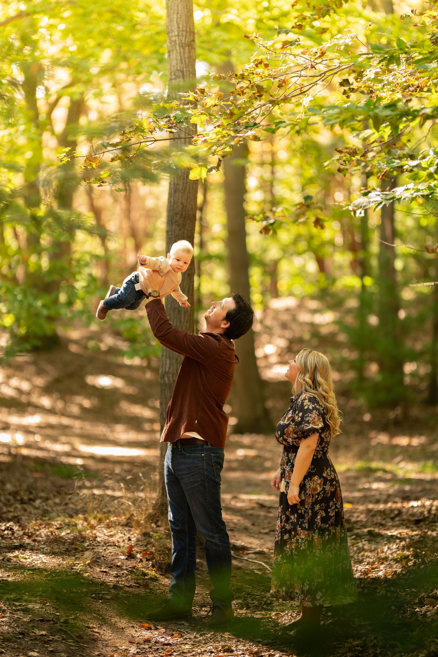 Family enjoying autumn forest walk, father lifting baby while mother watches among golden sunlit trees.