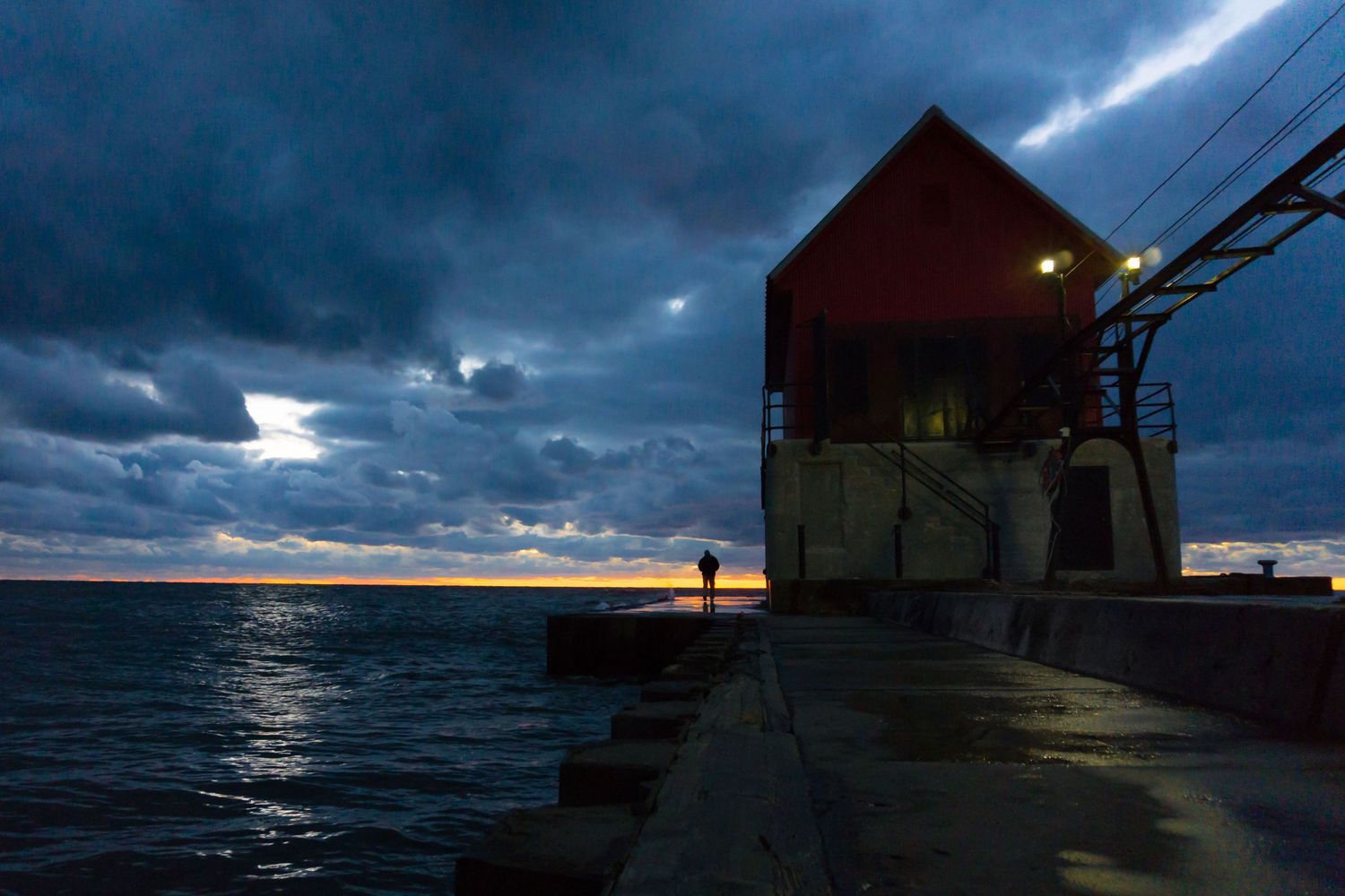 Lone figure stands beside a red harbor building at dusk under dramatic stormy clouds over the sea.