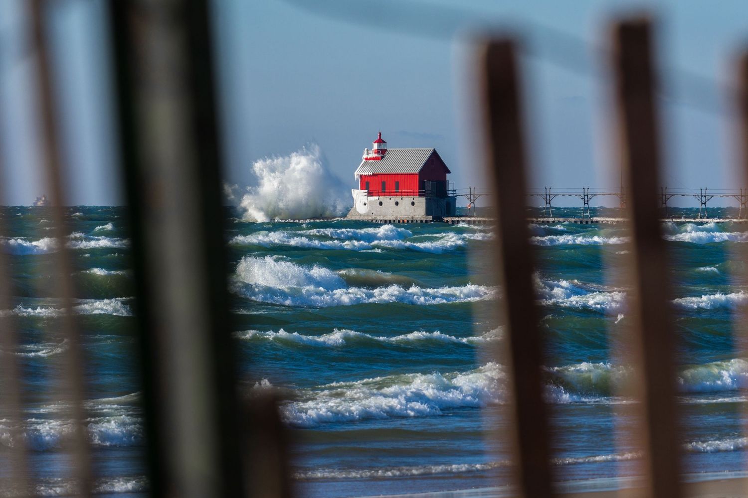 Red and blue lighthouse viewed through wooden fence slats amid crashing waves on a windy day.