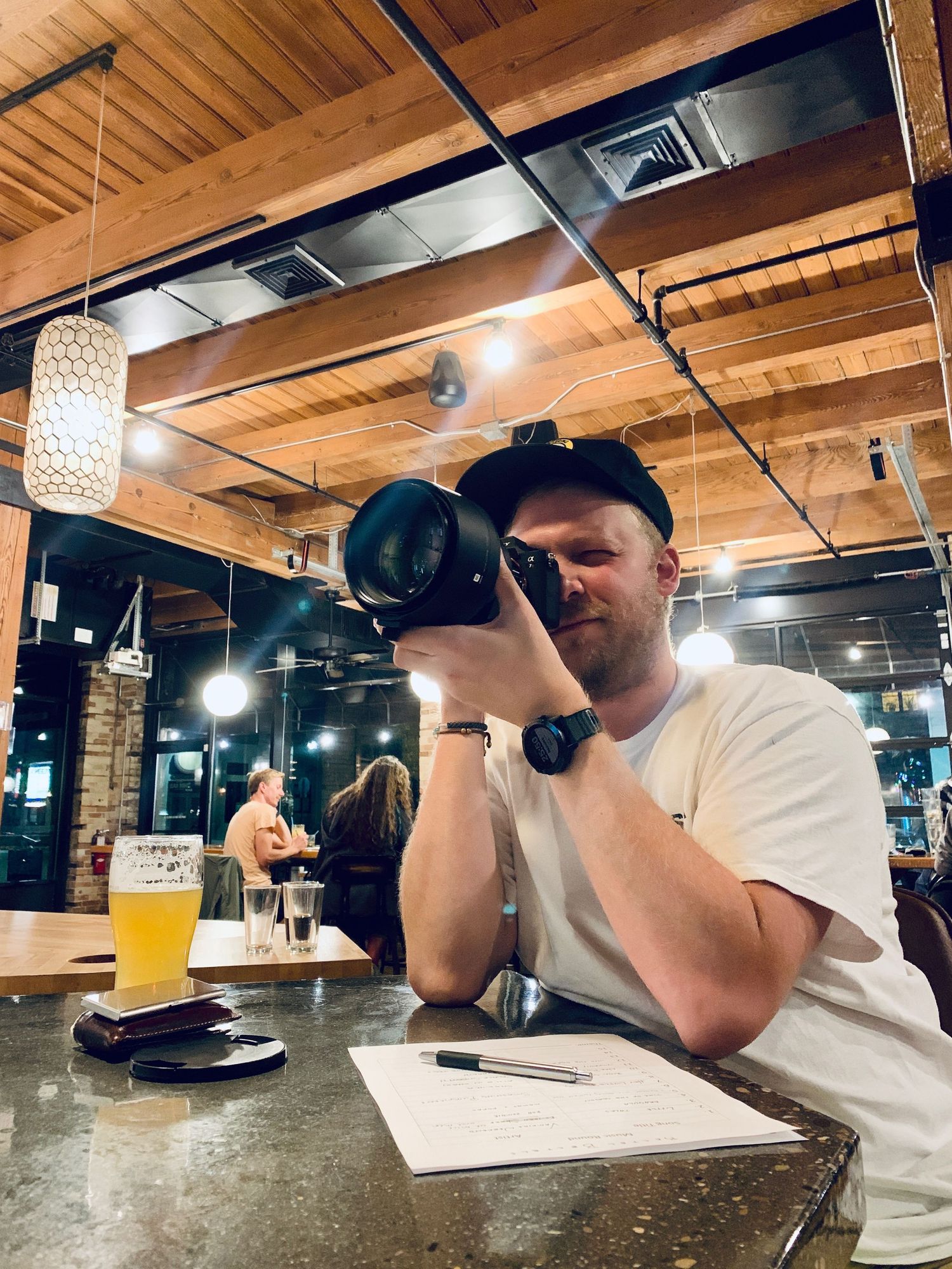 Man photographing with a large camera lens at a bar with a beer and notes nearby.