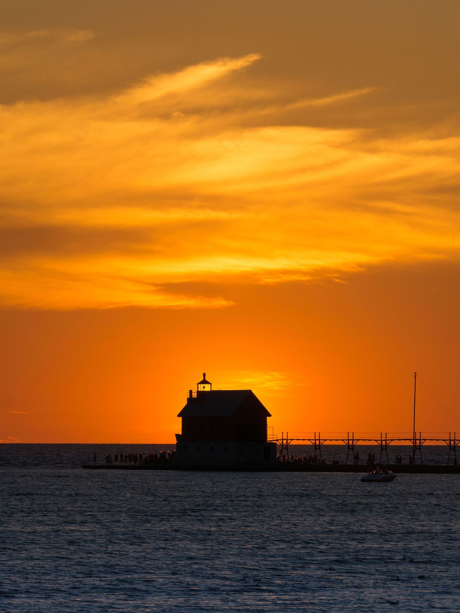 Silhouette of a lighthouse and pier against a vivid orange sunset sky reflecting on calm lake waters.