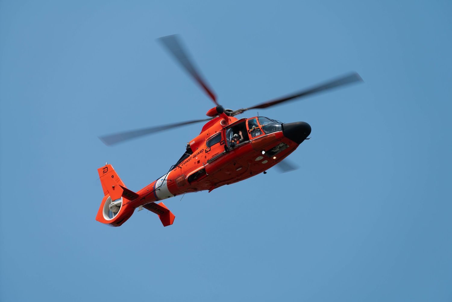 Bright orange US Coast Guard rescue helicopter flying against a clear blue sky.