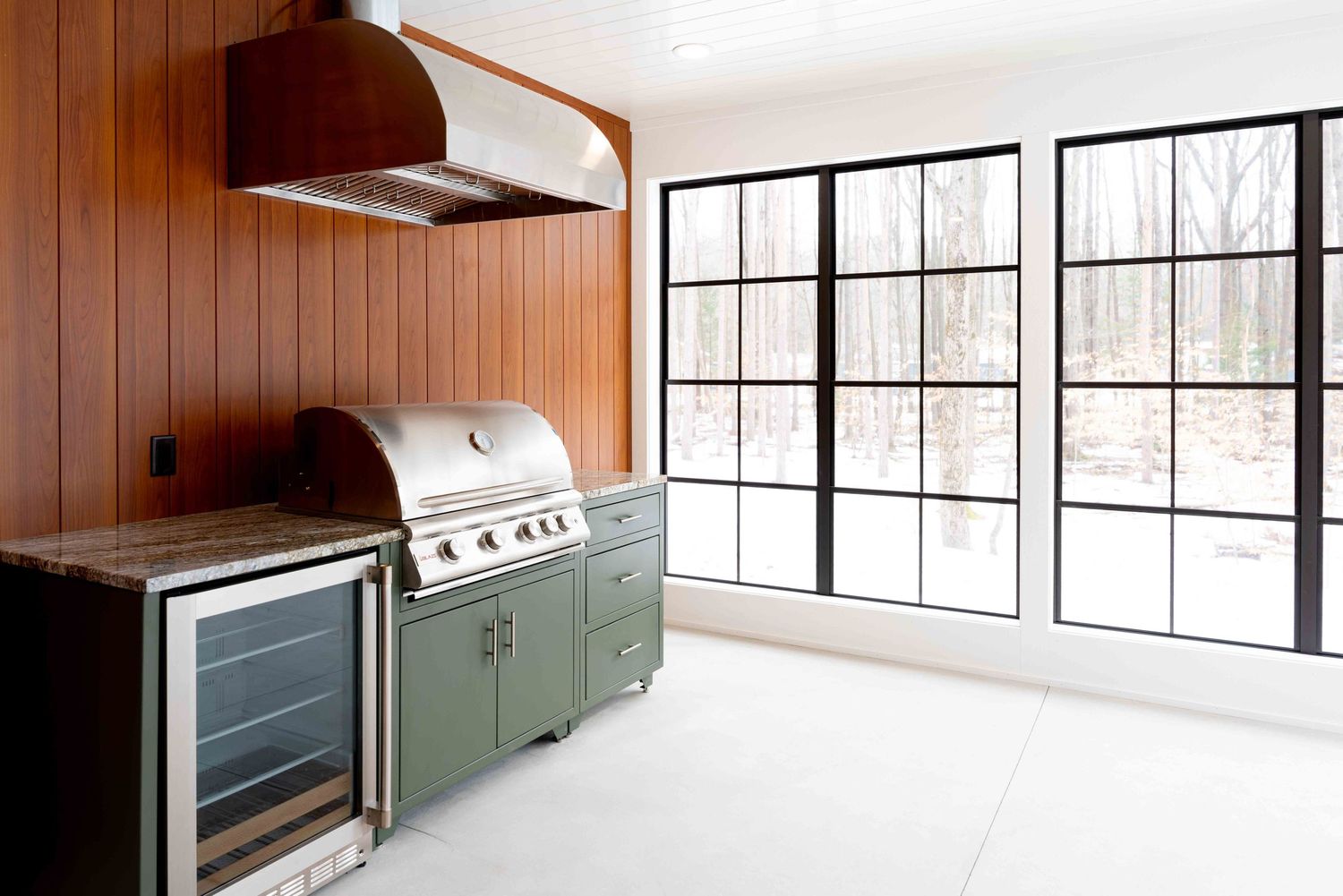 Outdoor kitchen with green cabinets, built-in grill, wine fridge, and black steel windows.