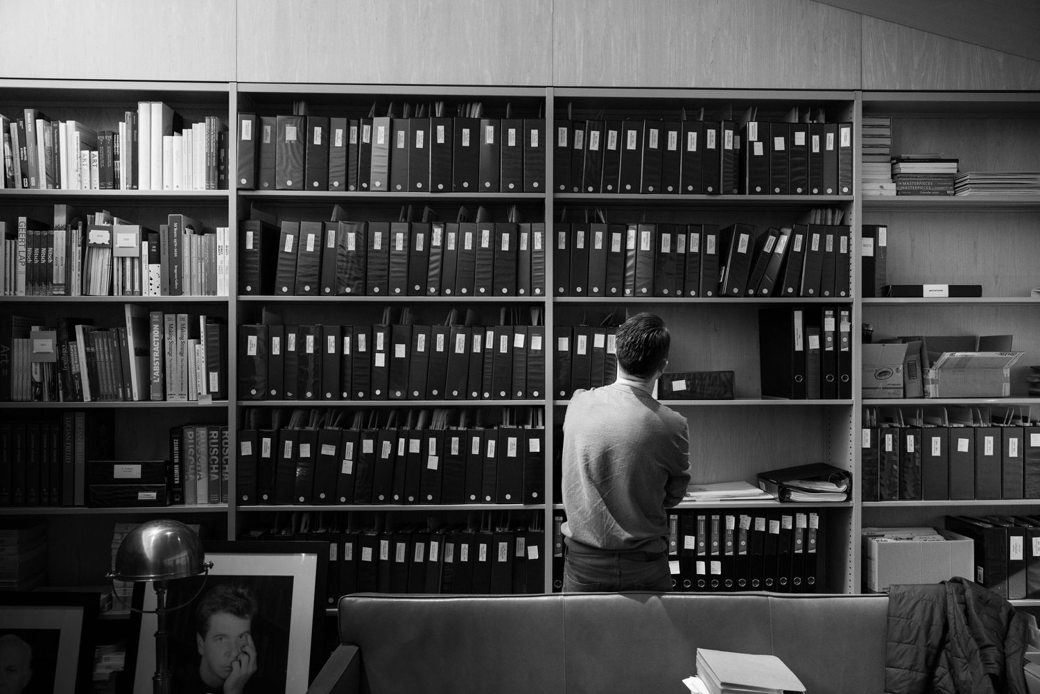 Man standing at bookshelves filled with binders and books in a library or archive, black and white.