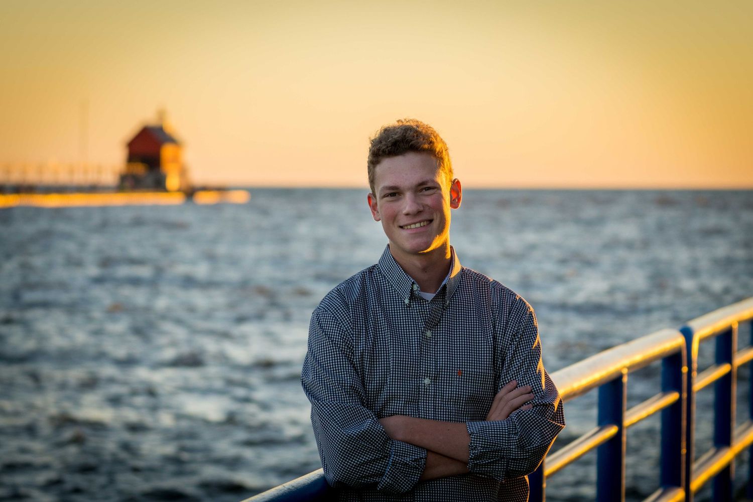 Smiling young man with crossed arms leaning on railing at sunset by lighthouse.