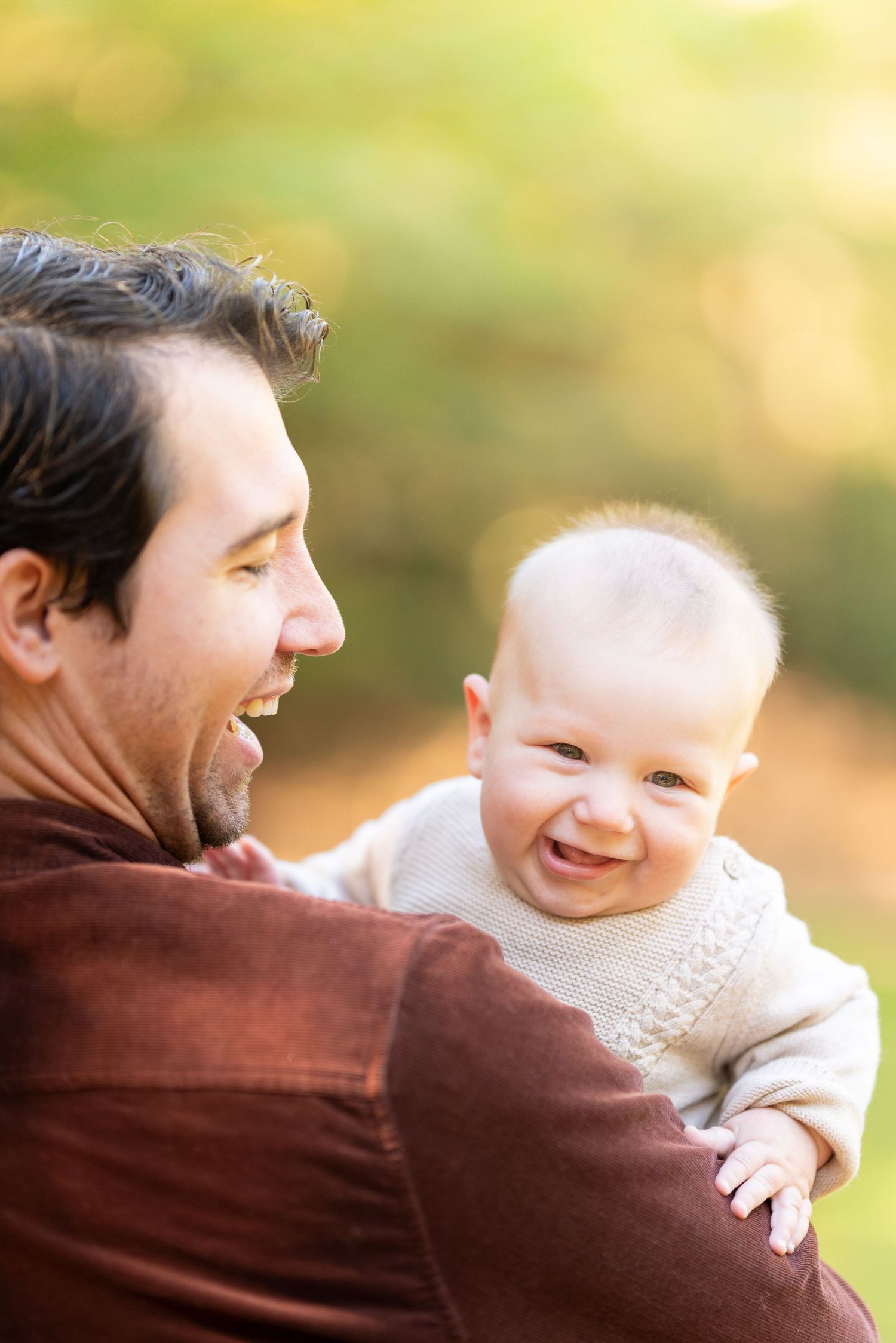 A laughing baby in a cream sweater smiles while being held by a bearded man outdoors in autumn.