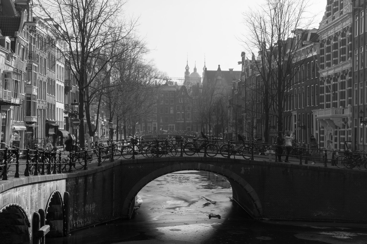 Black and white photograph of historic Amsterdam canal bridge with frozen water and city buildings.