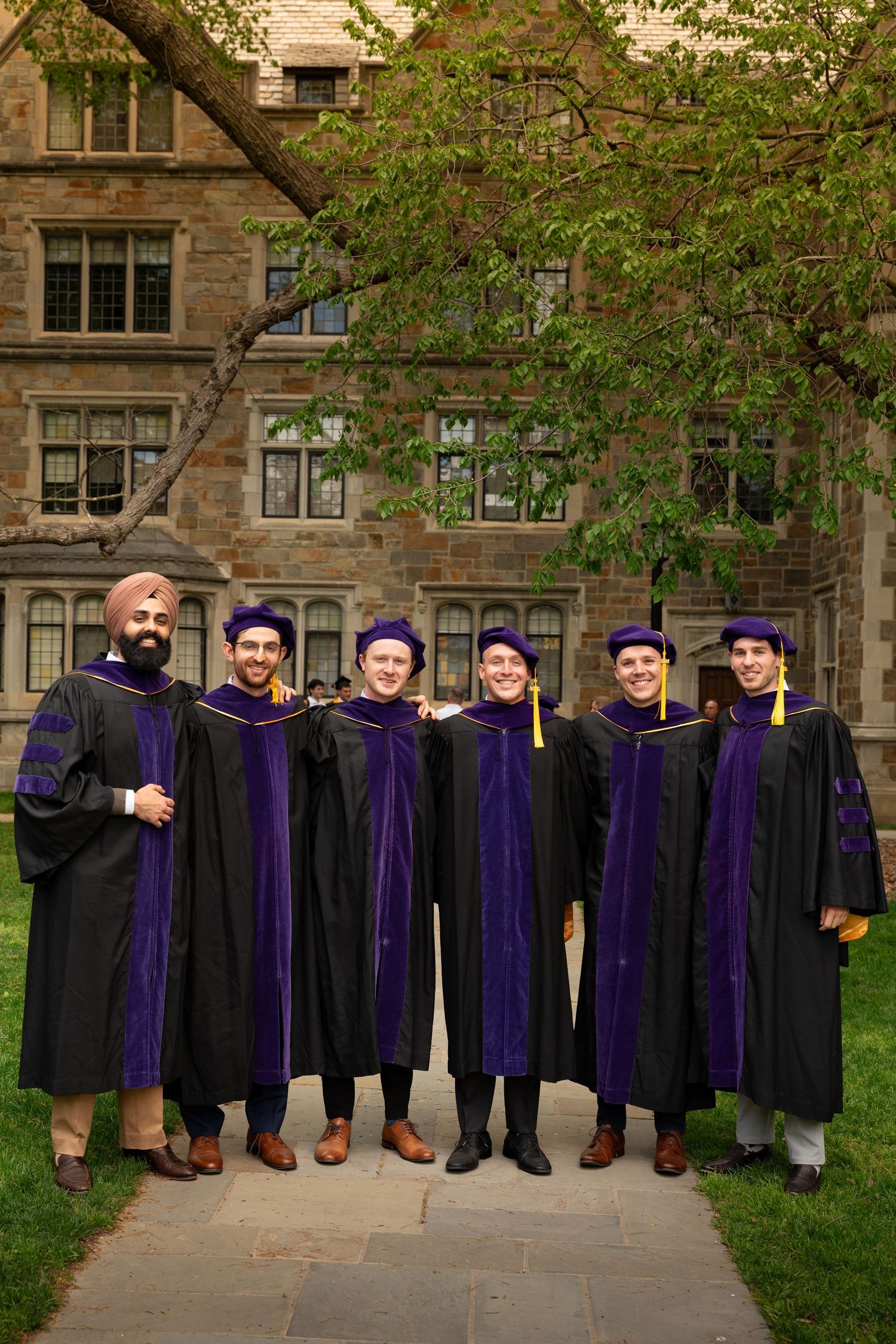 Group of six graduates in black and purple robes posing on campus pathway.