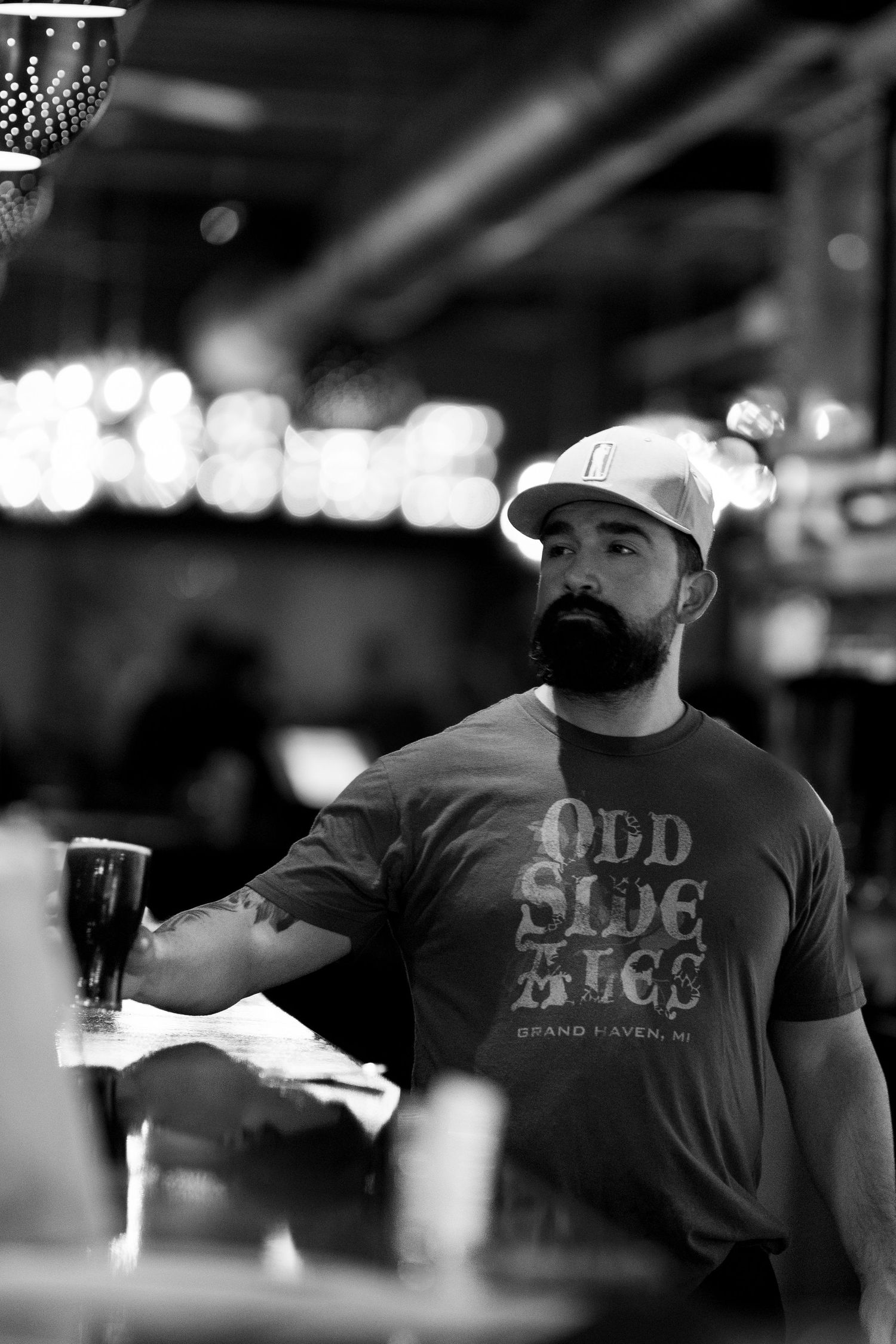 Bearded man in Odd Side Ales shirt holding a beer glass at a bar in black and white.